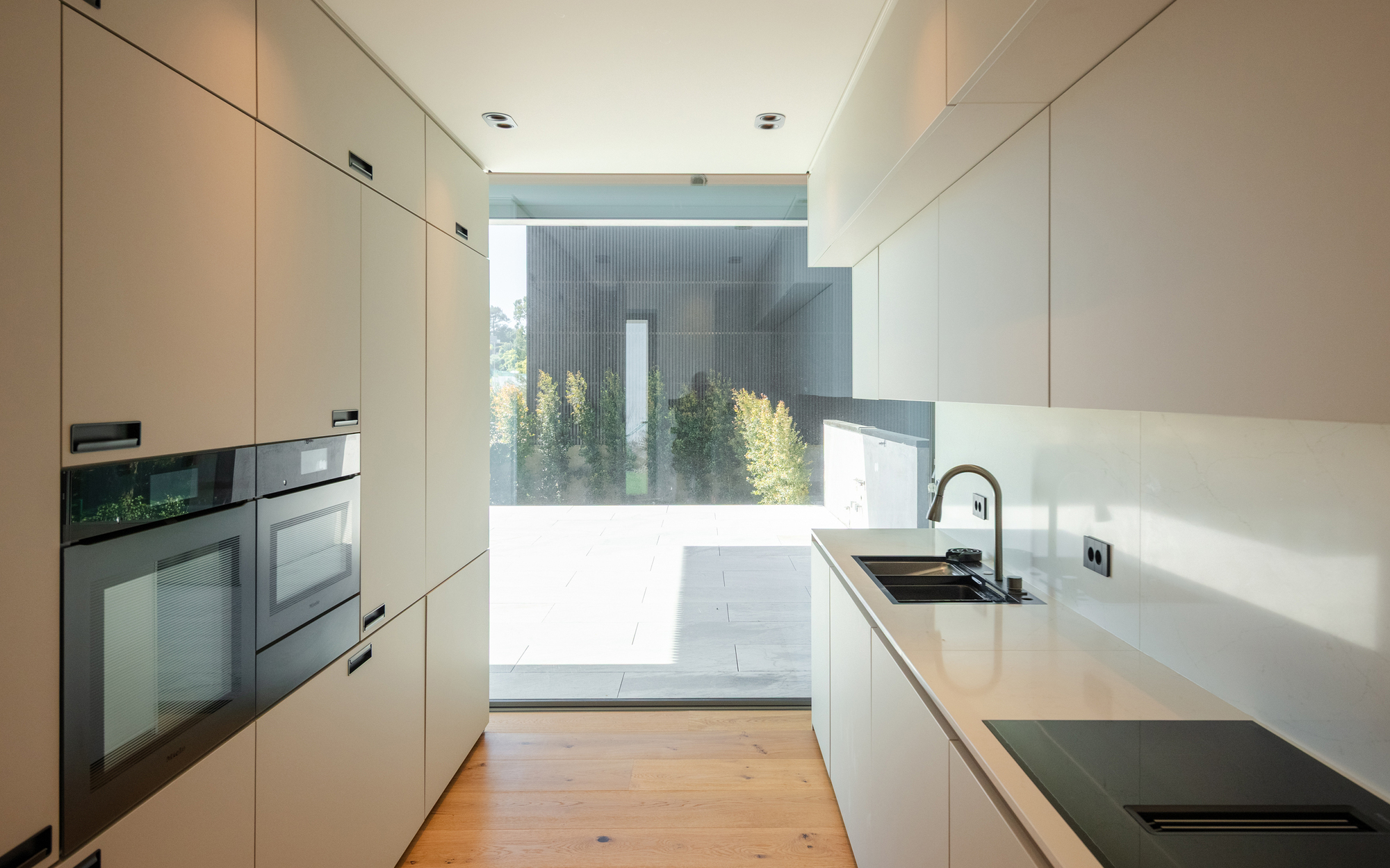 Narrow galley kitchen with white cabinets, built-in ovens and oak floor, opening through a glass wall to a sunlit paved terrace