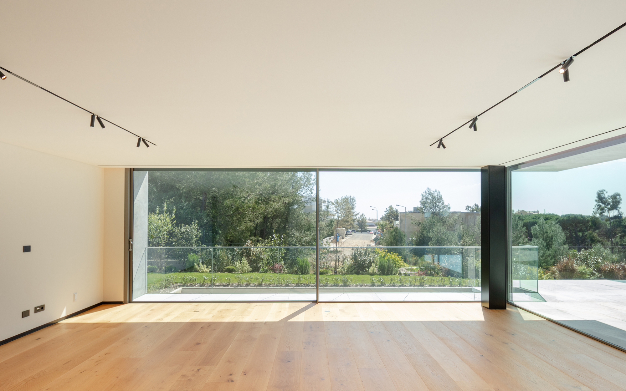Living room with floor-to-ceiling sliding glass opening onto a glass-balustrade terrace and garden, with track lights above and oak floor in sunlight
