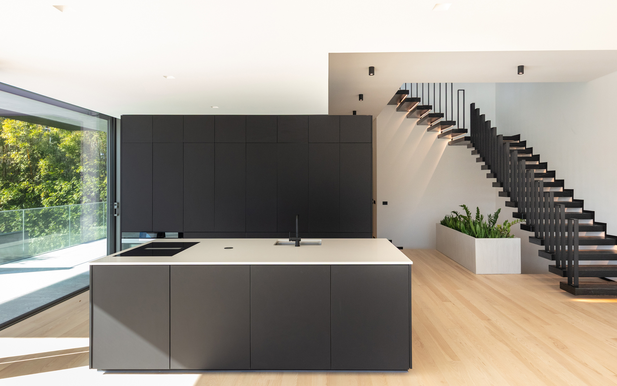 Black kitchen island and tall cabinet wall facing a floating black steel staircase, with a planter and garden light from the left