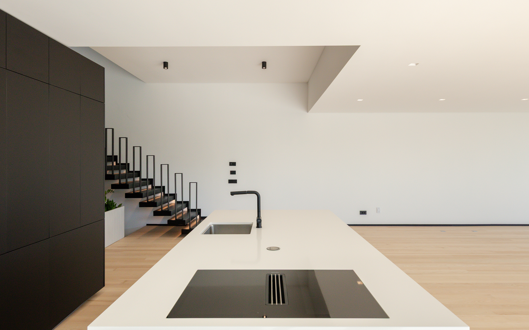 Long white island with sink and flush cooktop, black cabinet wall at left and a steel stair rising behind in soft daylight