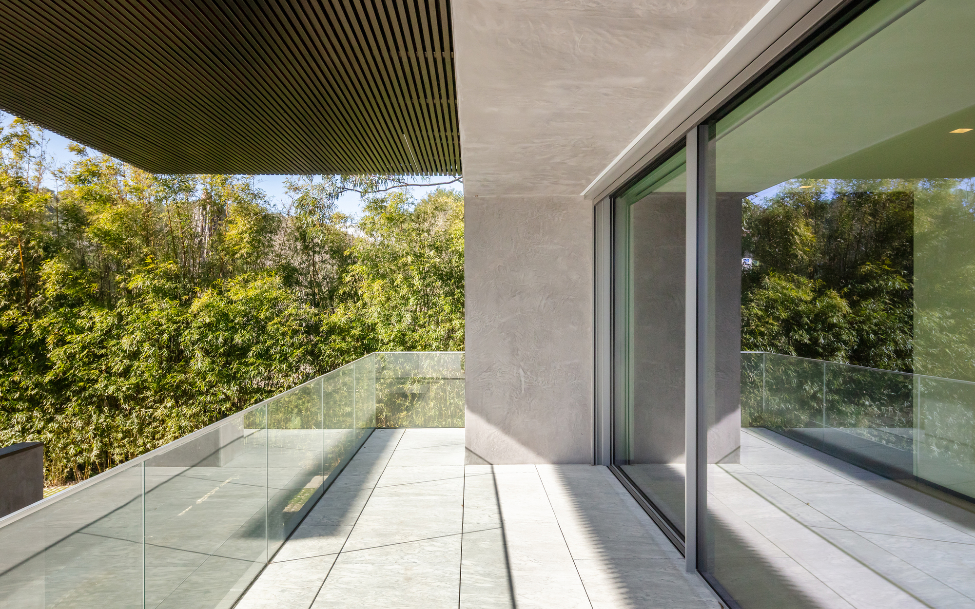 Covered terrace with slatted timber soffit, glass balustrade and stone-tile floor, framed by dense green trees in bright sun