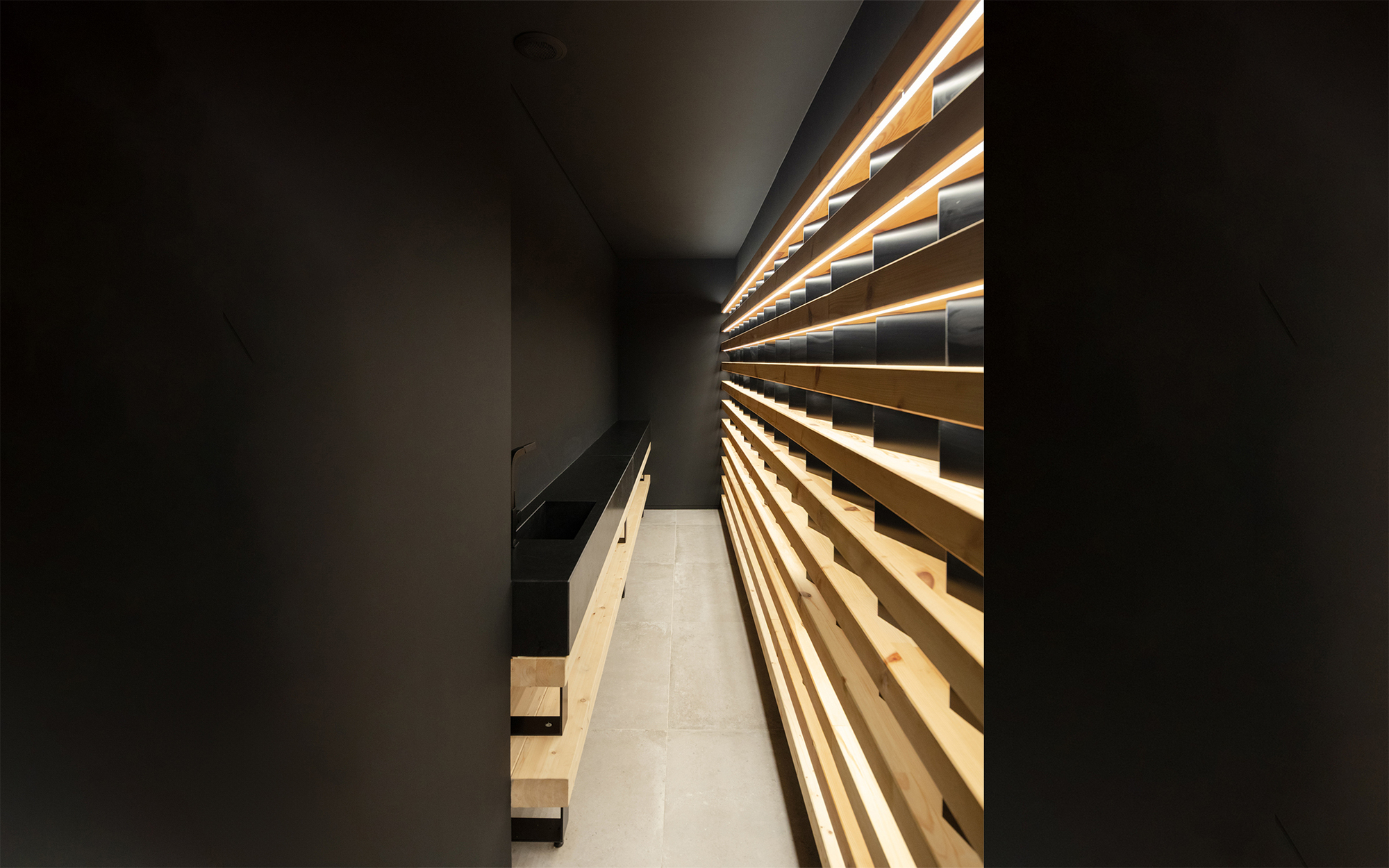 Narrow dark corridor lined with stacked timber slats lit from within, with a black basin and timber base at left