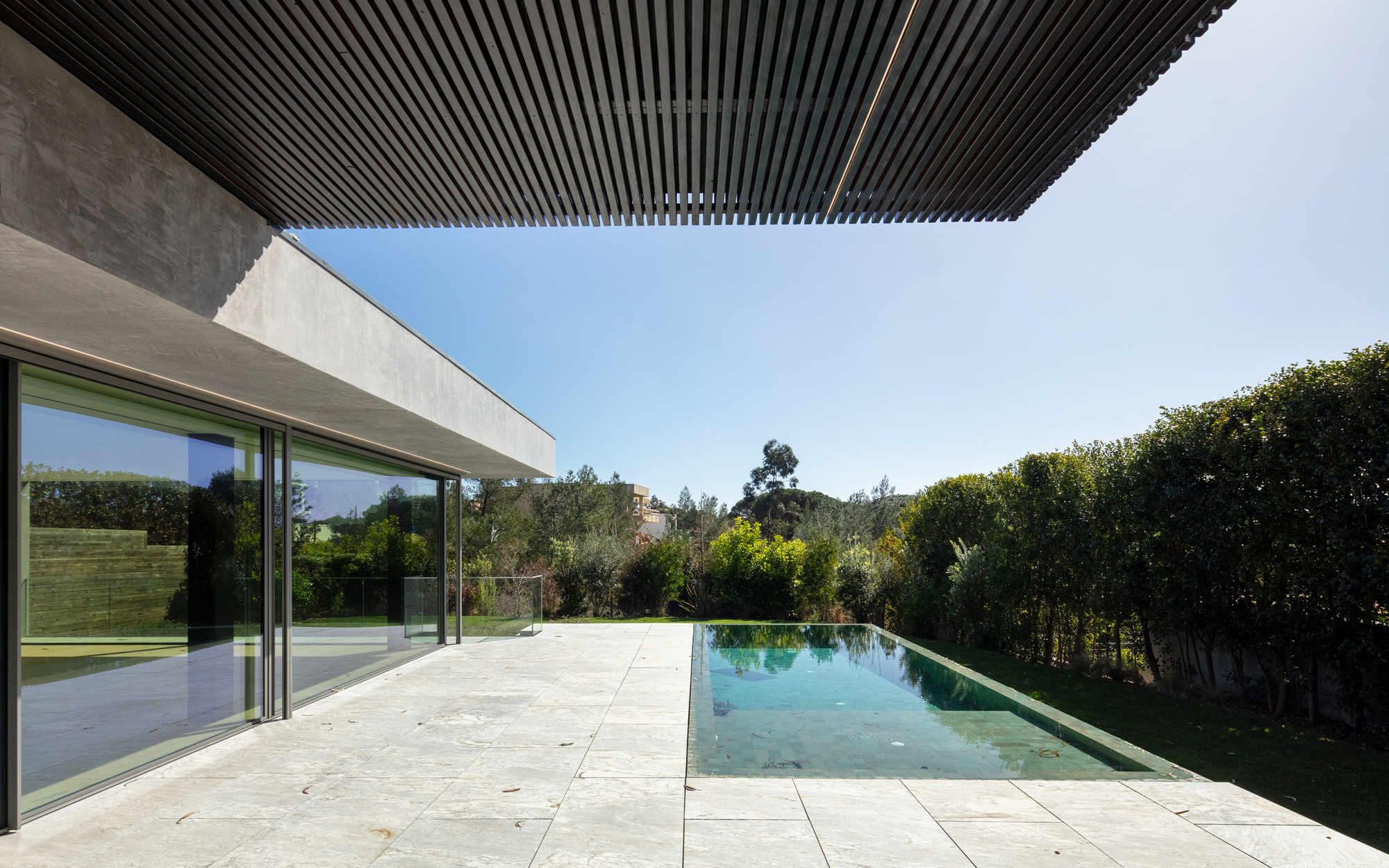 Slatted timber soffit cantilevering over a stone terrace and infinity pool, with a hedge and trees under clear blue sky