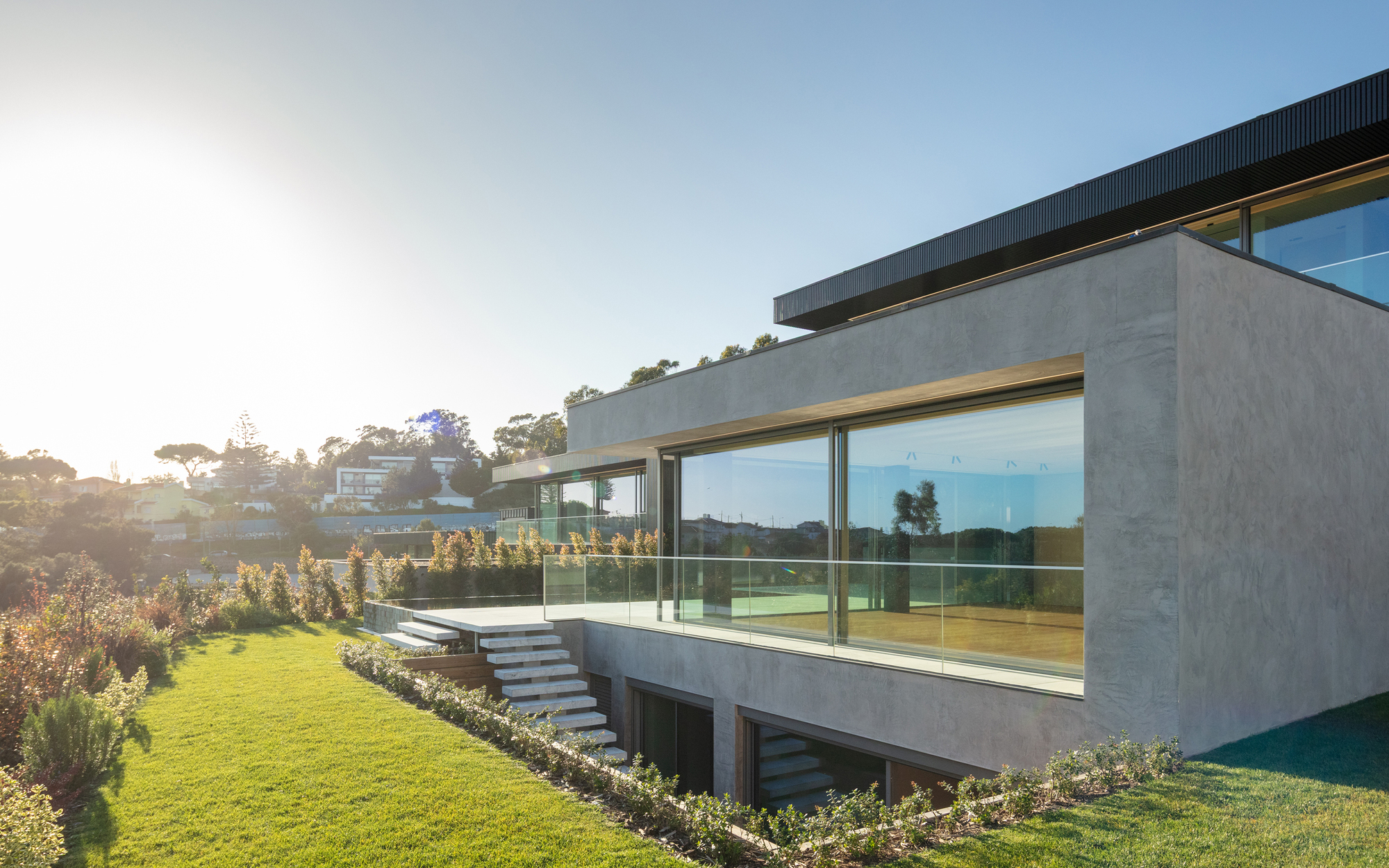 Concrete-rendered volume with glass balustrade and floating stone steps, seen across a lawn in low backlit sun