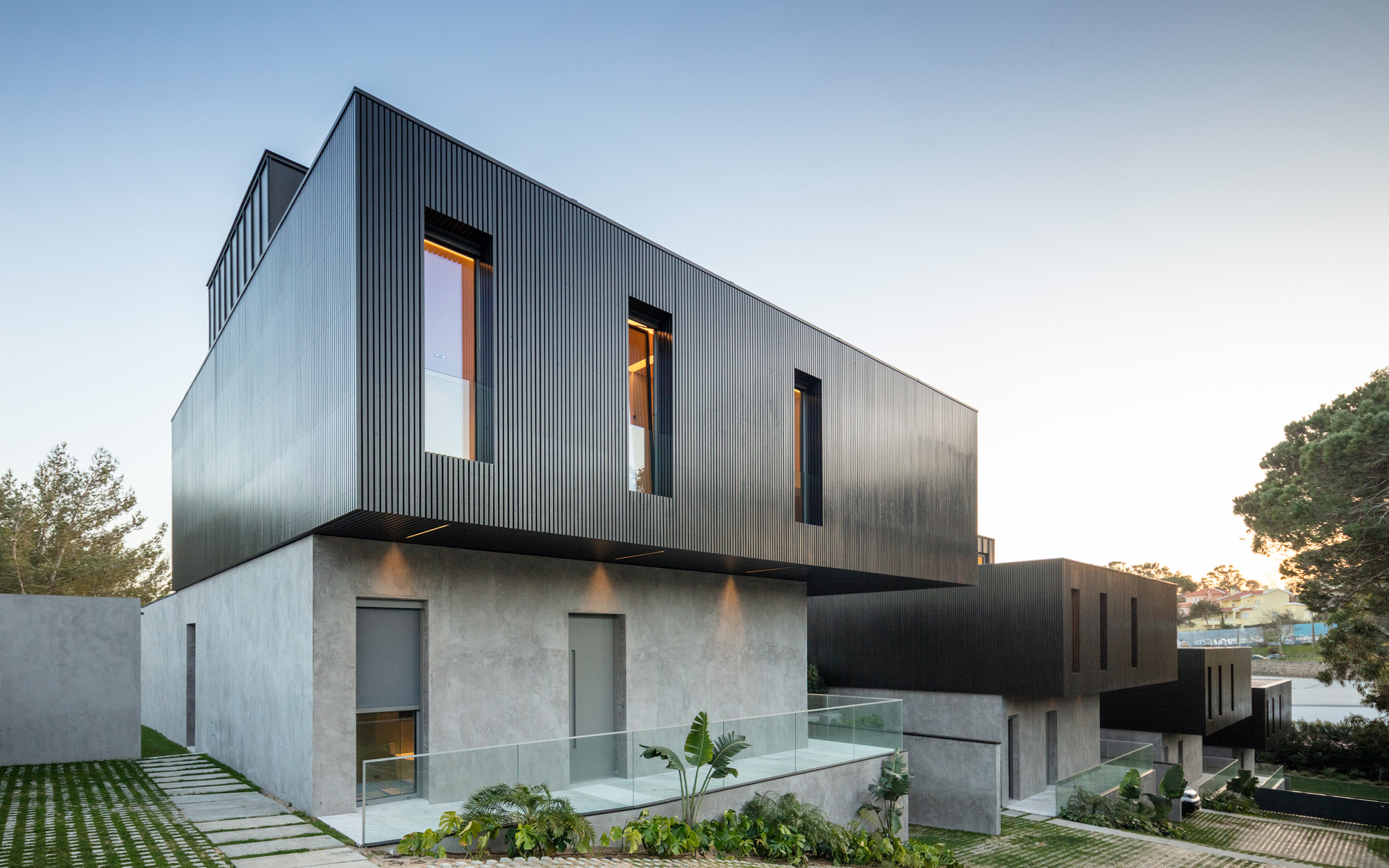 Black ribbed metal upper volume stacked on a grey concrete base with glass guardrail and palm plants at dusk