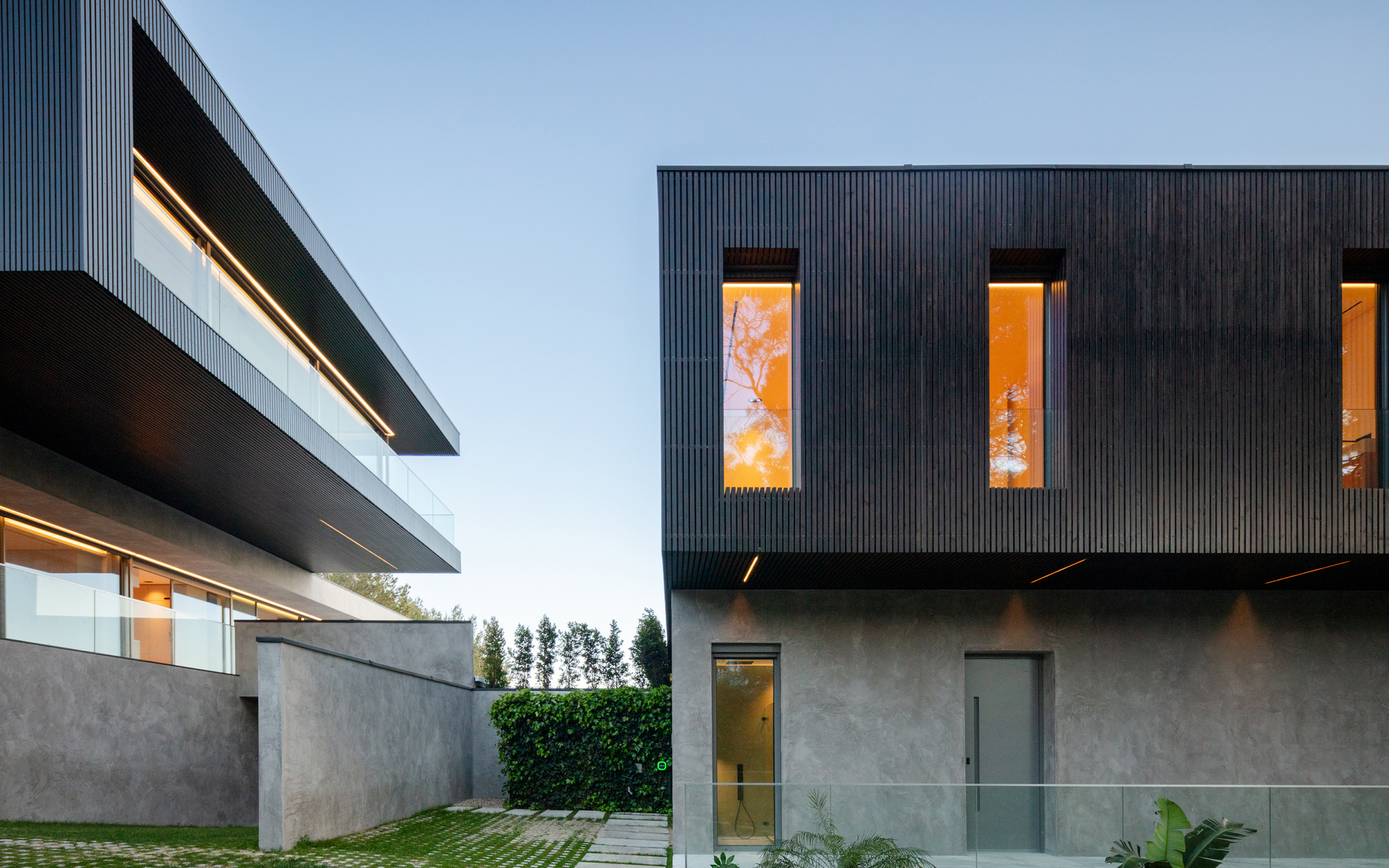 Two facades with dark vertical timber cladding and tall glowing amber windows, a hedge between them at dusk