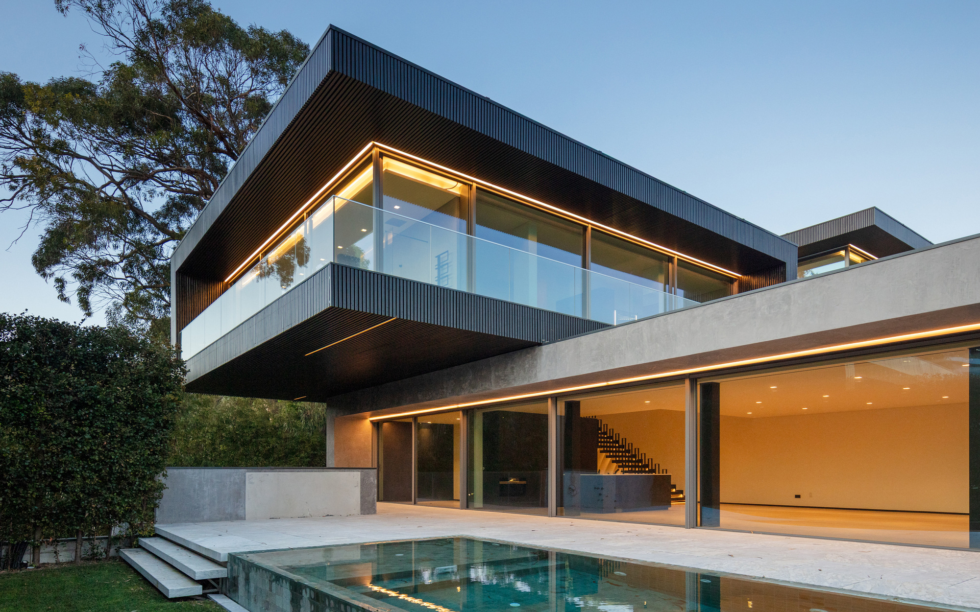 Cantilevered dark upper floor with glowing edge lighting above a glazed living space and a pool at dusk