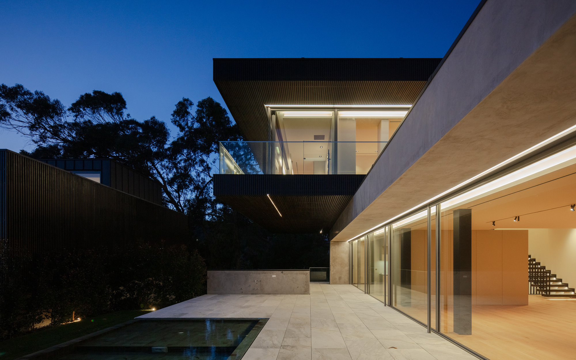 Dusk view of the cantilevered upper volume, glazed ground floor and stone terrace beside a reflecting pool under eucalyptus trees