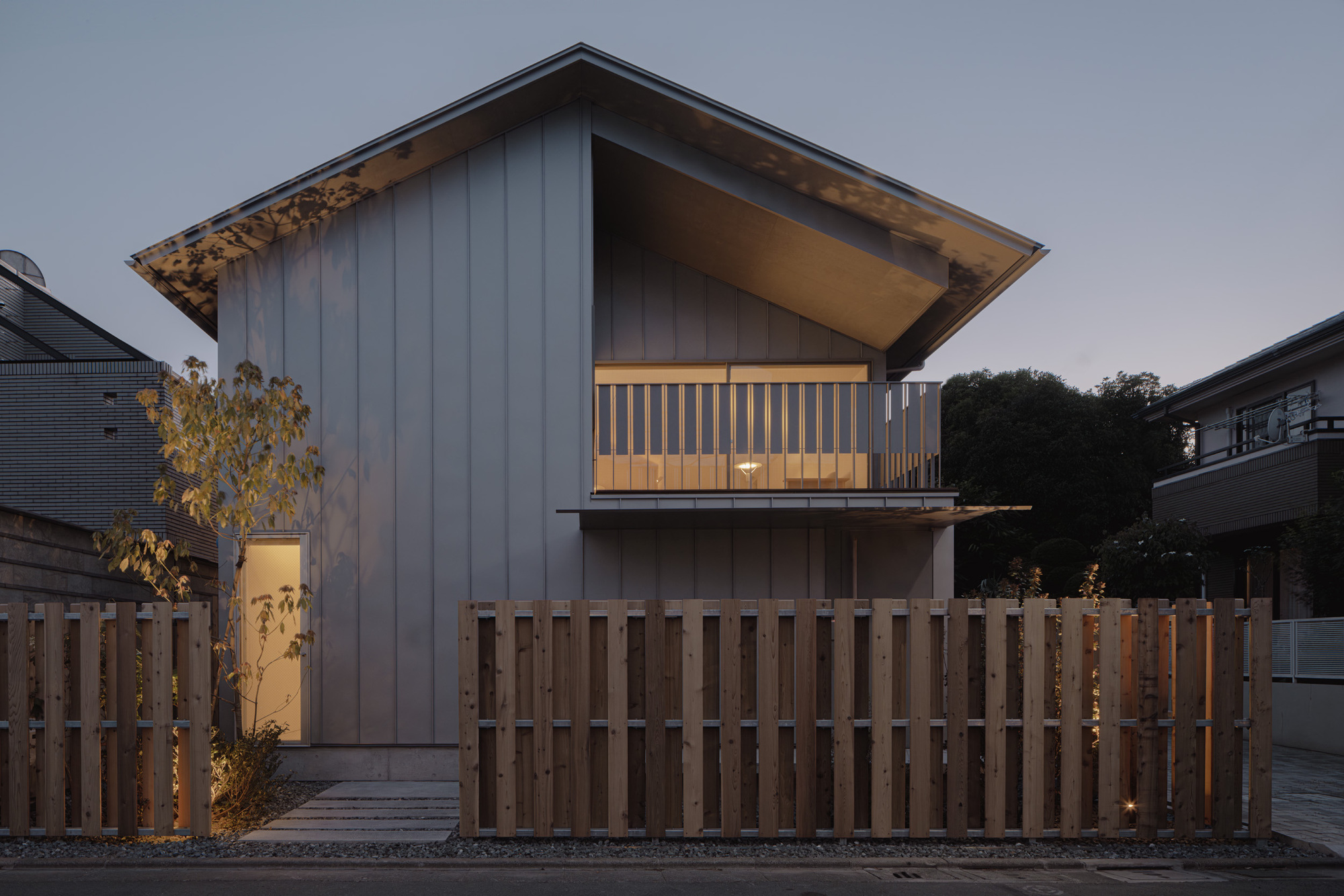 Gabled street facade clad in vertical grey panels behind a slatted timber fence, with a glowing balcony at dusk
