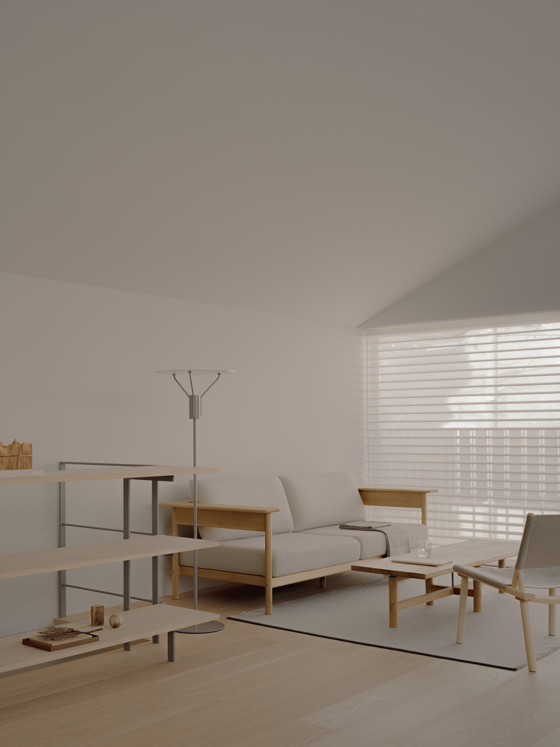 Living room with a wood-framed sofa, low table, open shelving and floor lamp beside a window covered by a sheer blind
