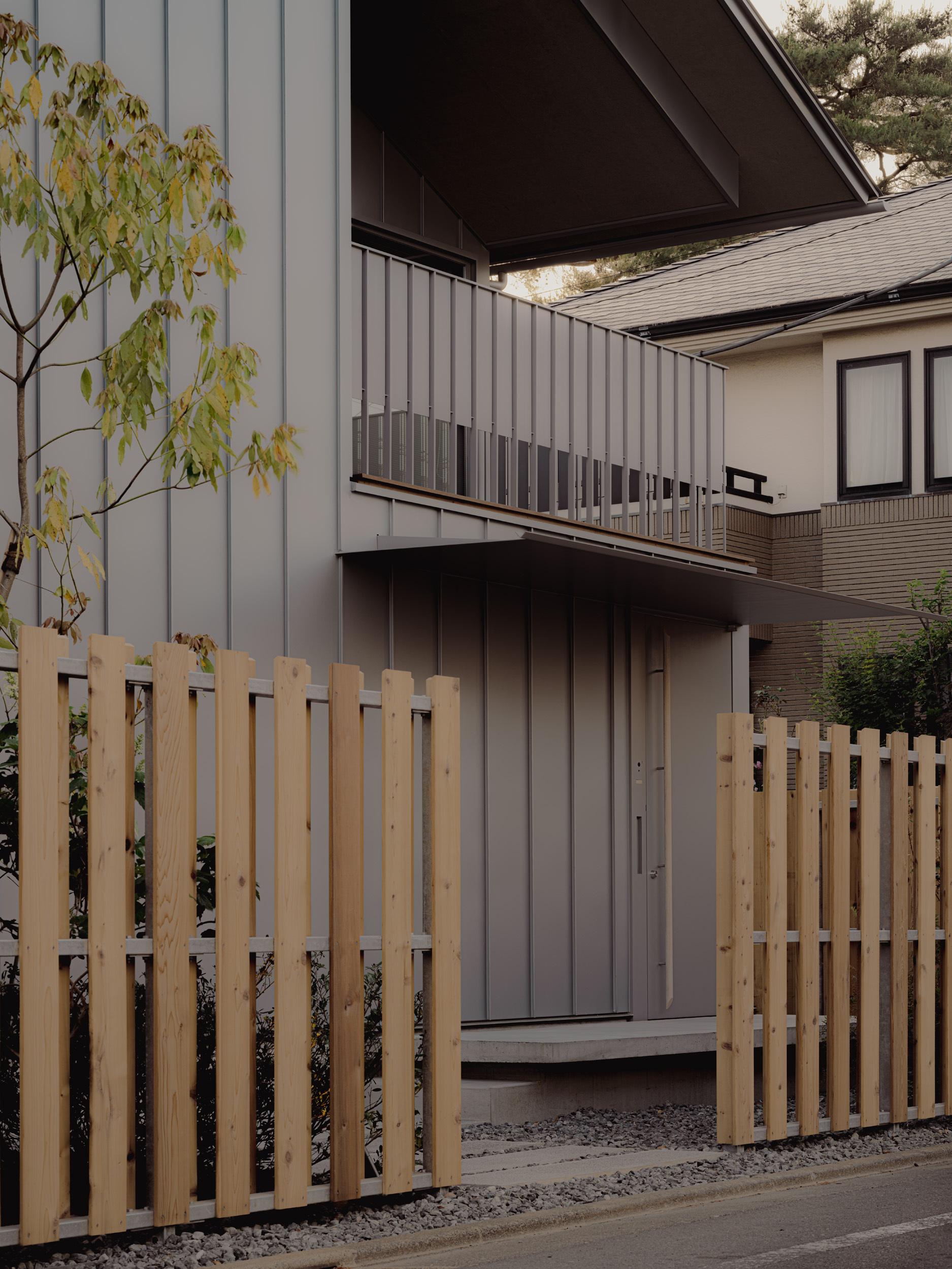Entrance front with a balcony of vertical balusters above a recessed door, framed by cedar fence panels and a small autumn tree