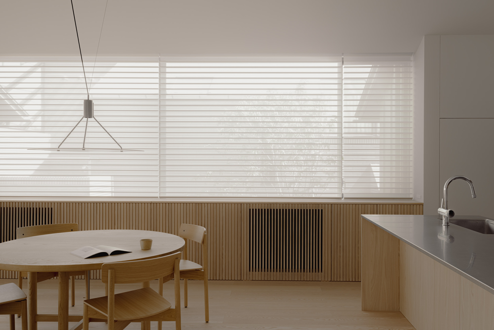Sheer blinds drawn over a wide window above slatted timber panelling, with a round table and steel kitchen counter in muted light