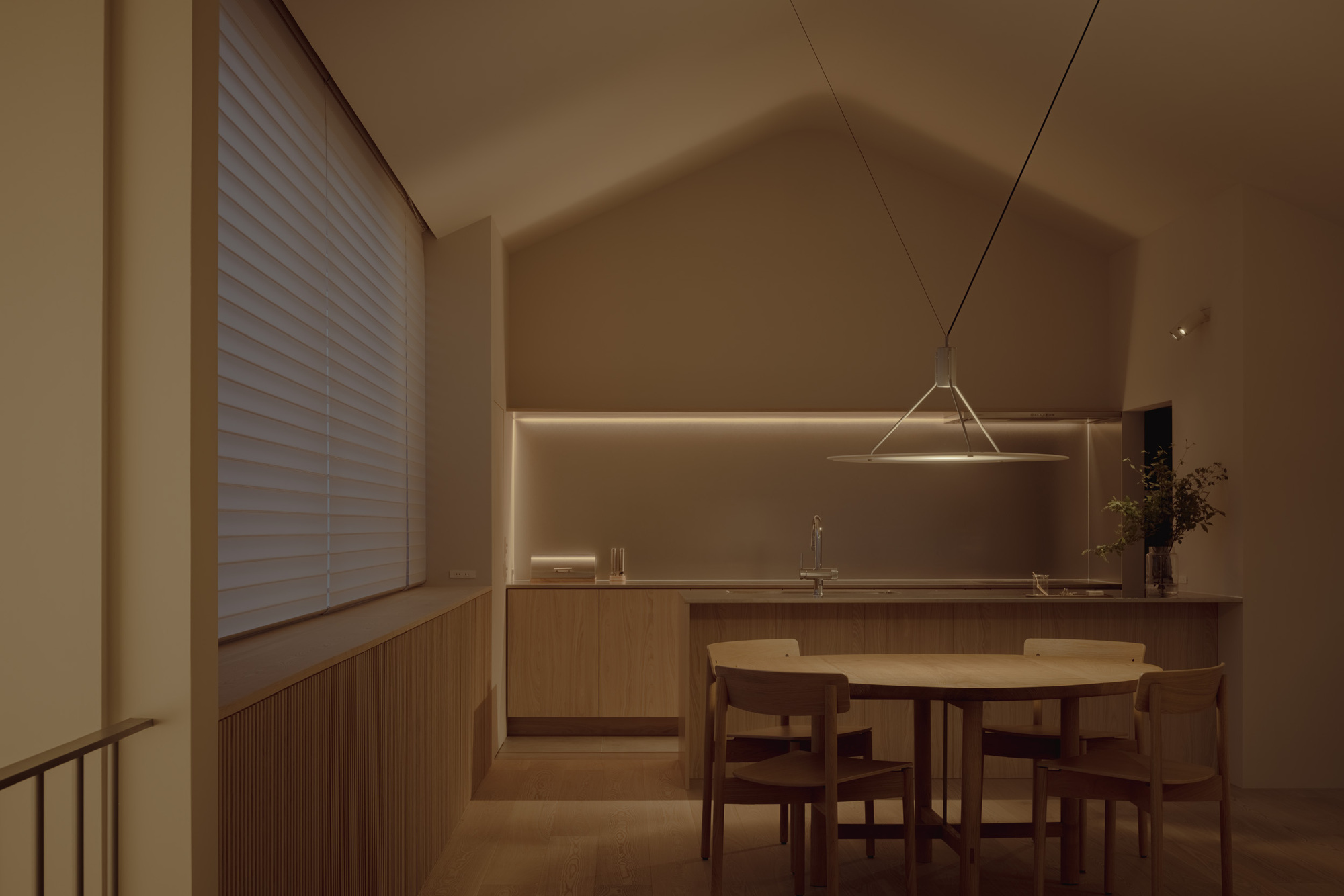 Kitchen and round oak dining table under a gabled ceiling, a disc pendant glowing with a slatted blind drawn at dusk