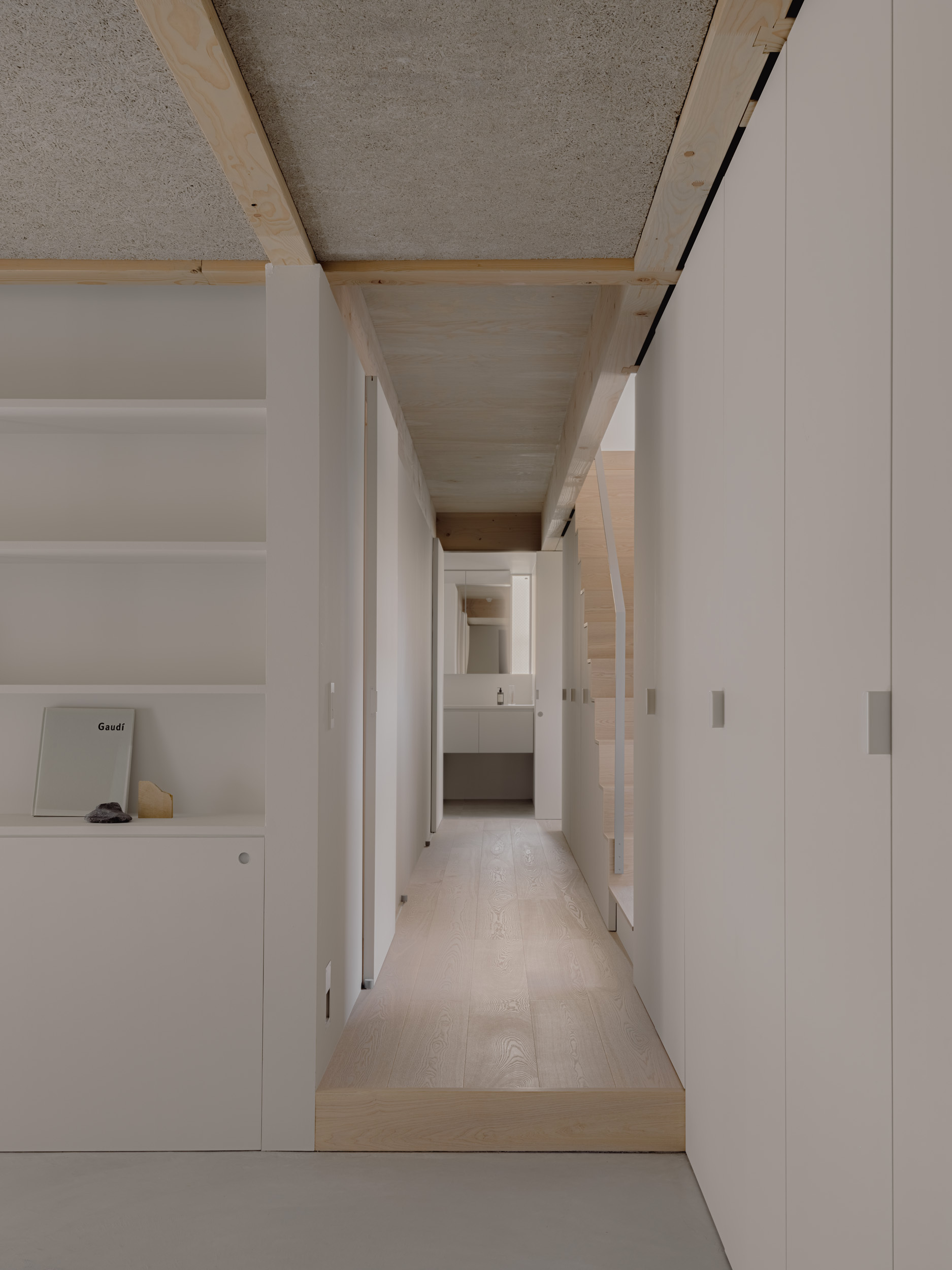 Narrow corridor with timber ceiling joists and hempcrete panels, white built-in cabinets on both sides leading to a bathroom in daylight