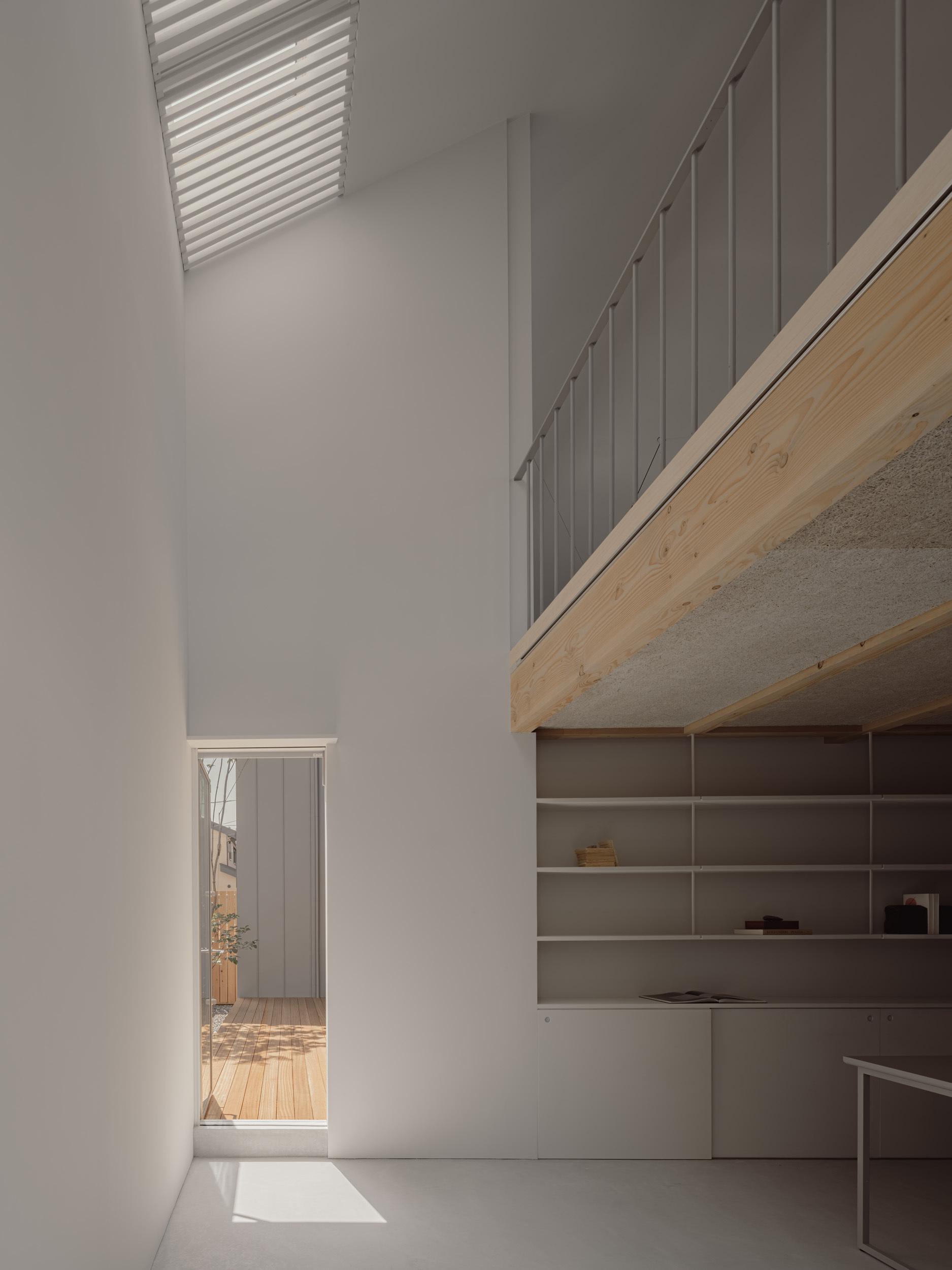 Double-height white room with a timber mezzanine edge, open shelving and a louvred rooflight casting a patch of sun on the floor