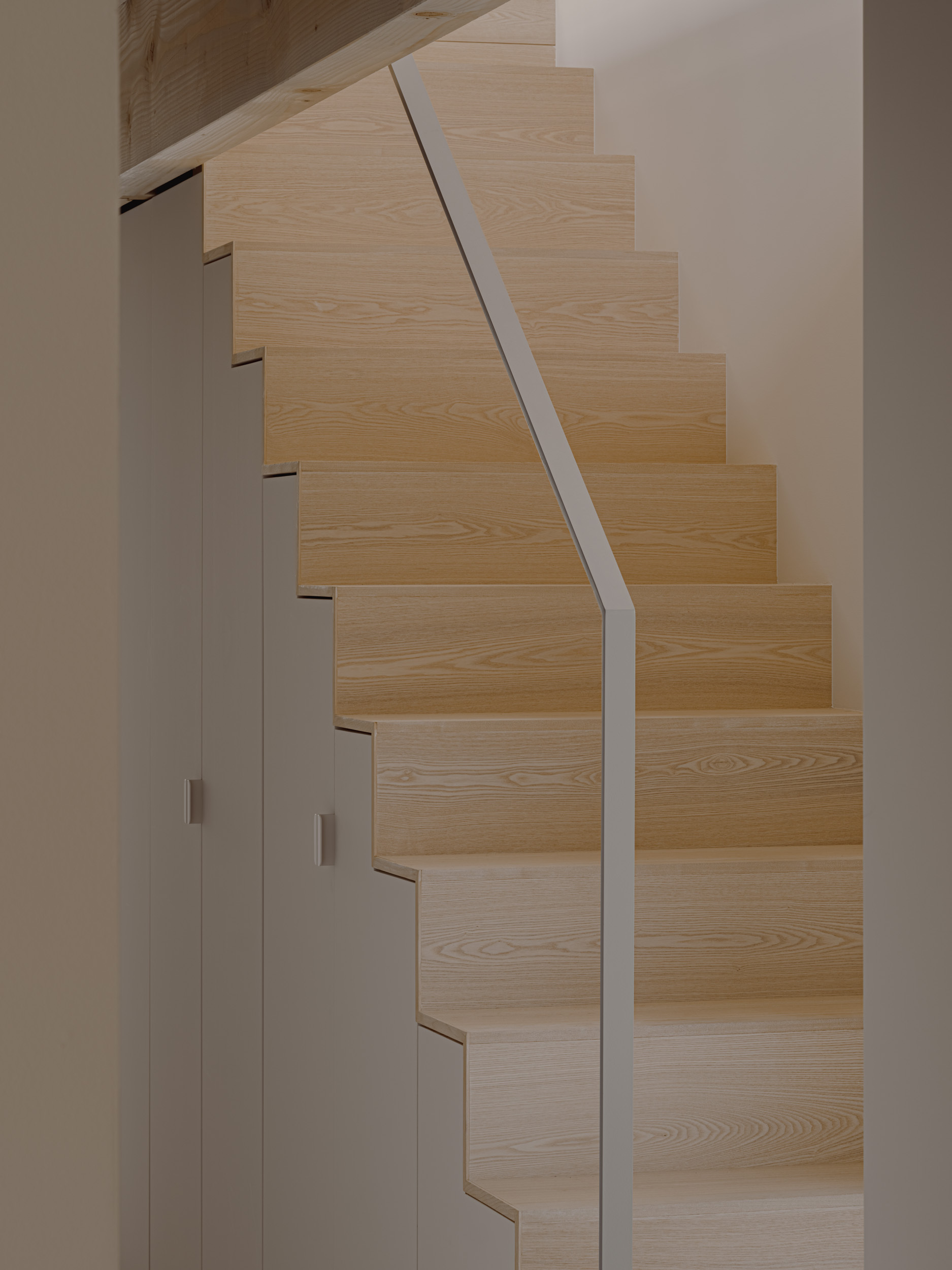 Pale ash staircase with a flat steel handrail and storage cupboards built into the side, lit by soft indoor light