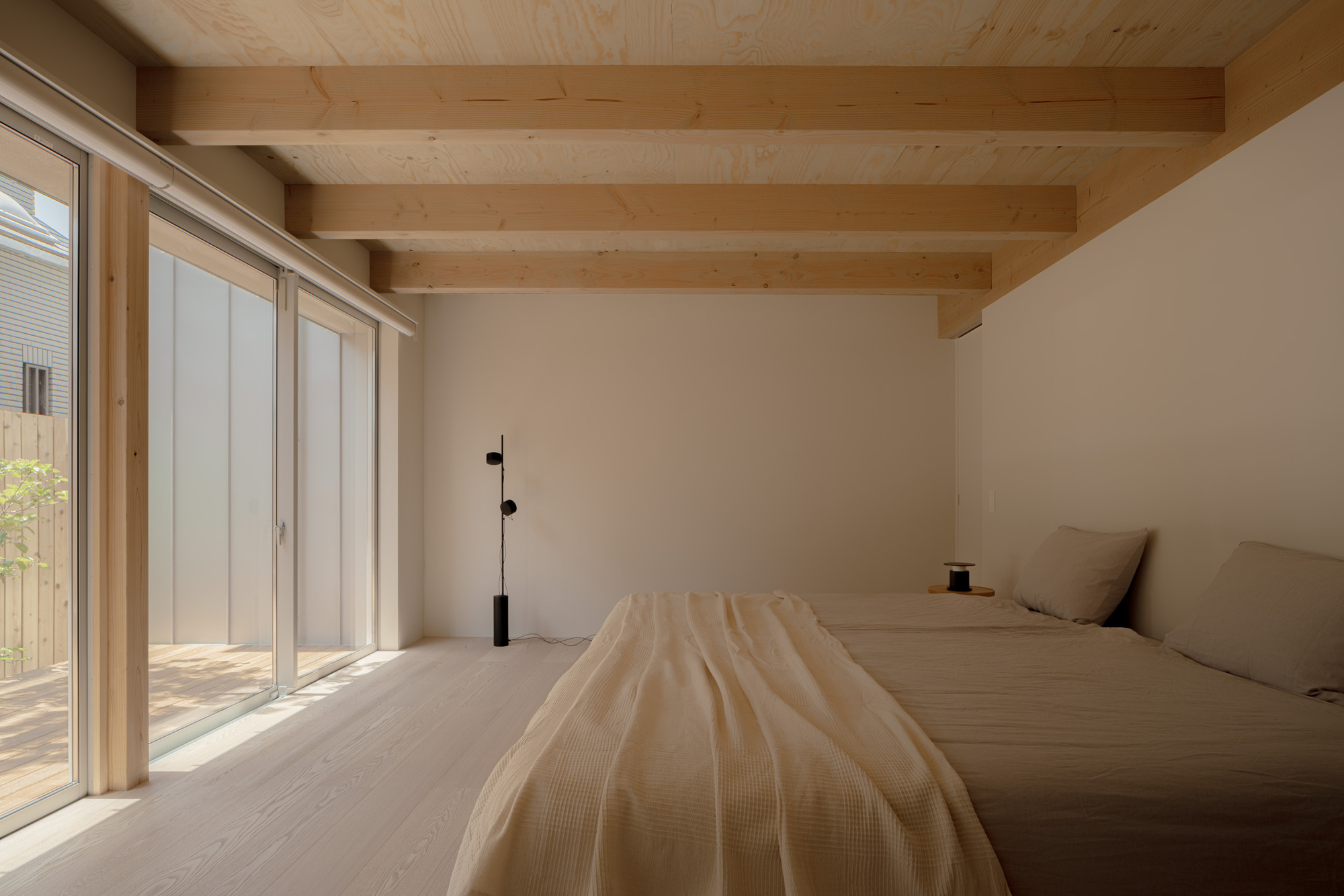 Bedroom with exposed timber ceiling beams and a low bed beside floor-to-ceiling glass doors opening to a sunlit wooden deck