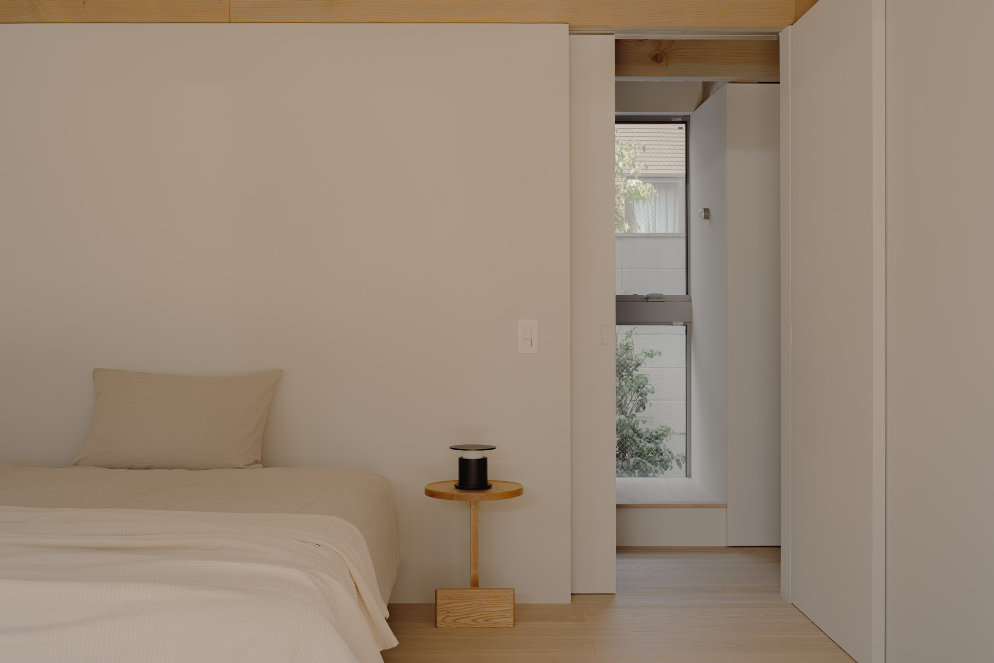 Bedside table with a black lamp beside the bed, and a sliding panel opening onto a narrow window with greenery outside