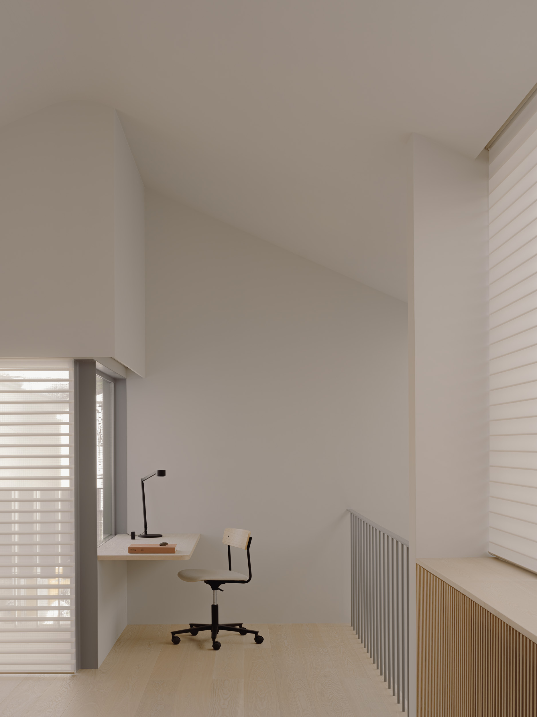 Loft study nook with a built-in desk and swivel chair under a sloped ceiling, filtered by sheer horizontal blinds