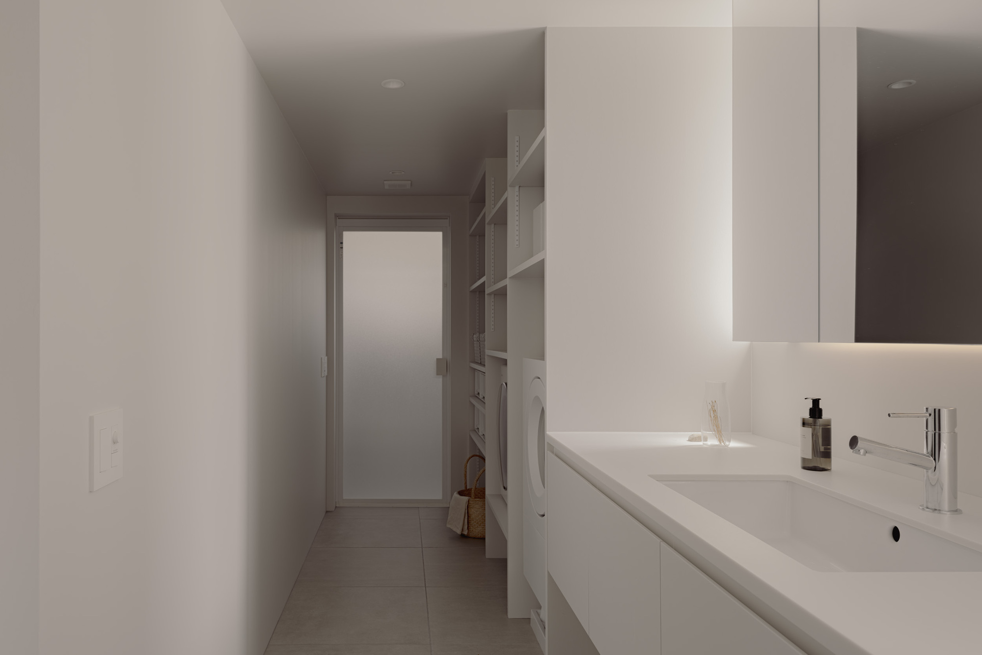 Narrow utility corridor with a white vanity and basin, open shelving, and a frosted glass door at the far end in soft light