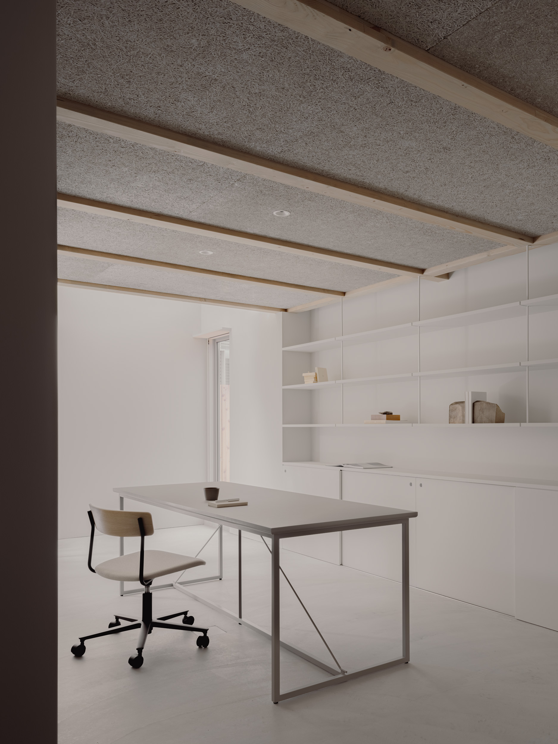 Work room with a steel-framed table and wooden chair beneath a wood-wool ceiling crossed by timber beams, with white shelving behind