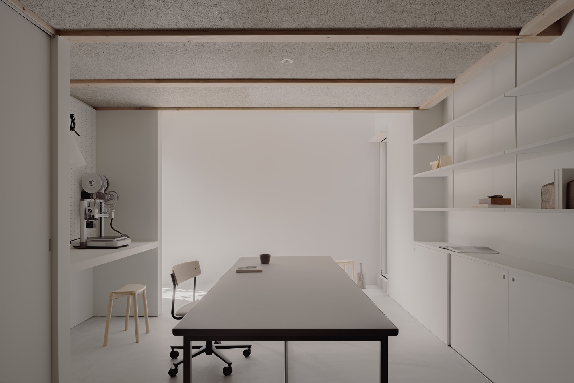 Long work table facing a white wall, with an espresso machine on a built-in counter and shelves under the beamed ceiling