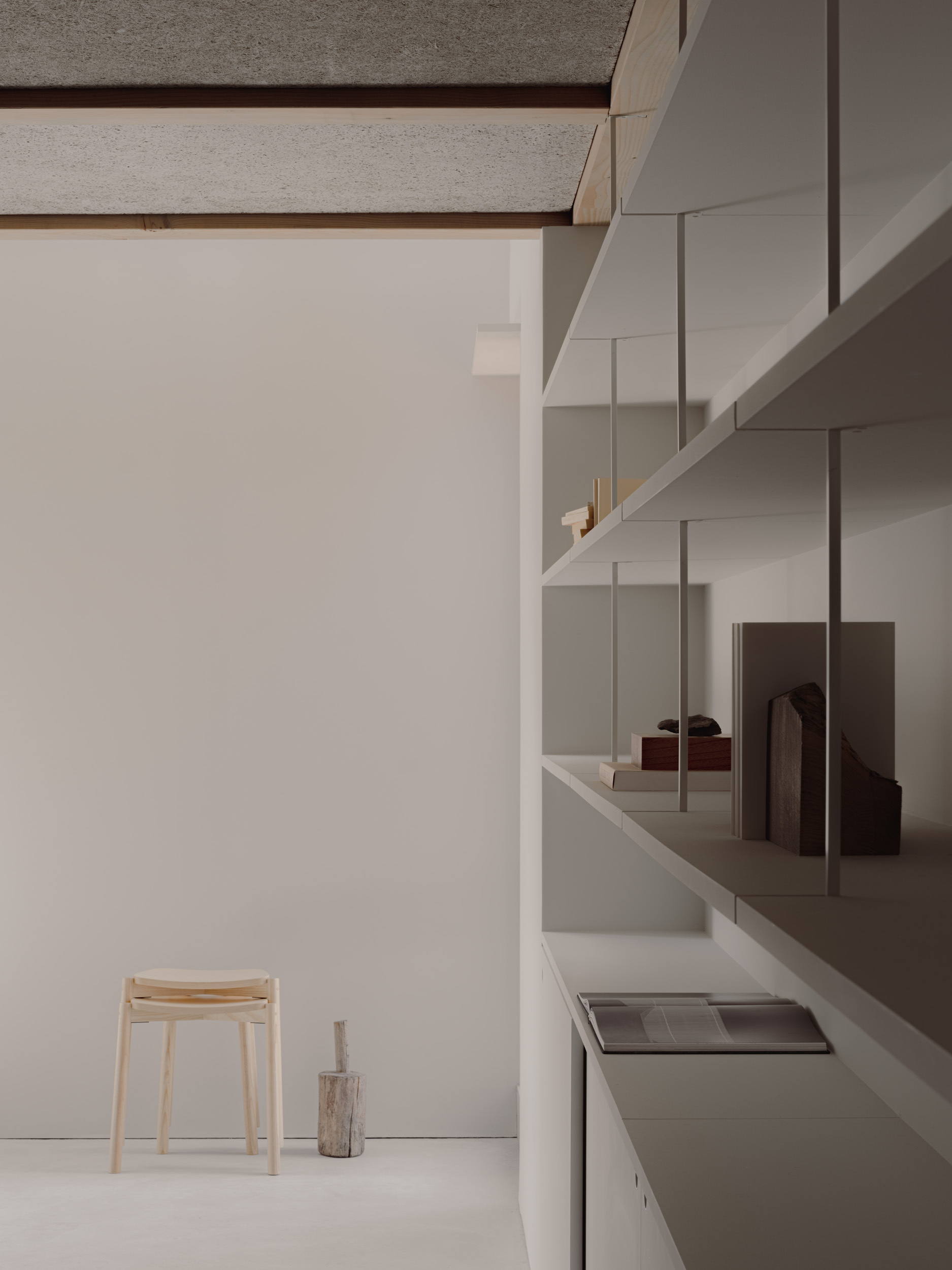 Wall-hung shelving with books and objects beside a wooden stool and log offcut, under a pale textured ceiling in soft light