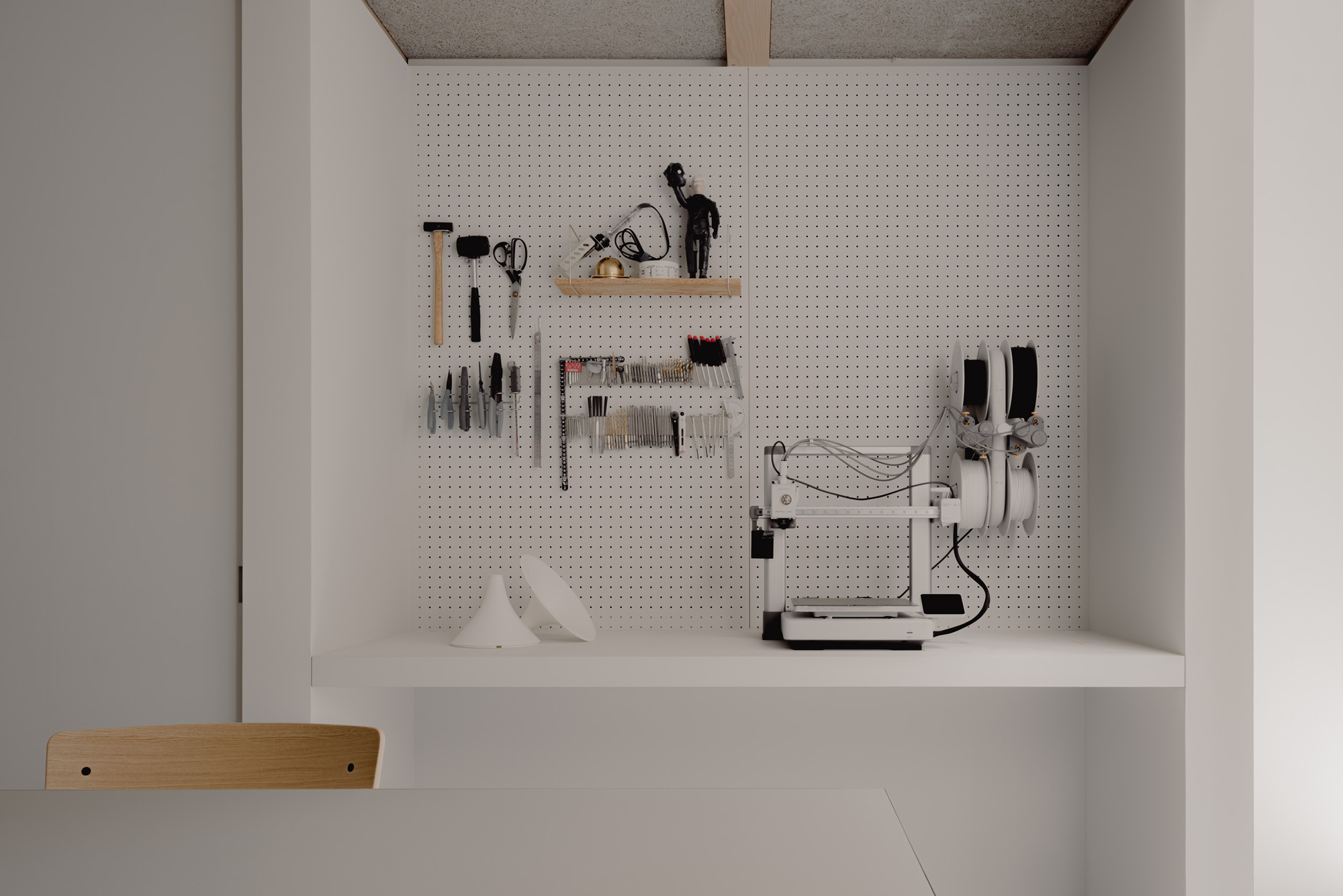 Pegboard workshop alcove with hanging tools, a small 3D printer and filament spools above a built-in white desk, a wooden chair in front