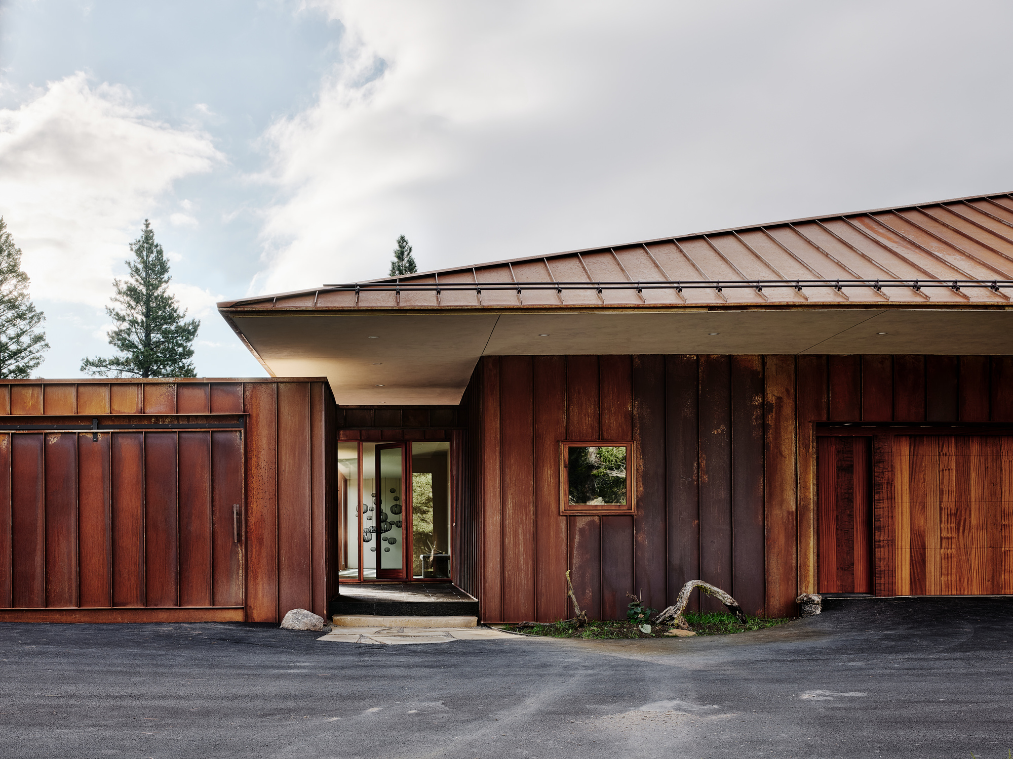 Entry with a deep canopy overhang, weathered steel panel walls and a small square window above a gravel forecourt, under passing clouds