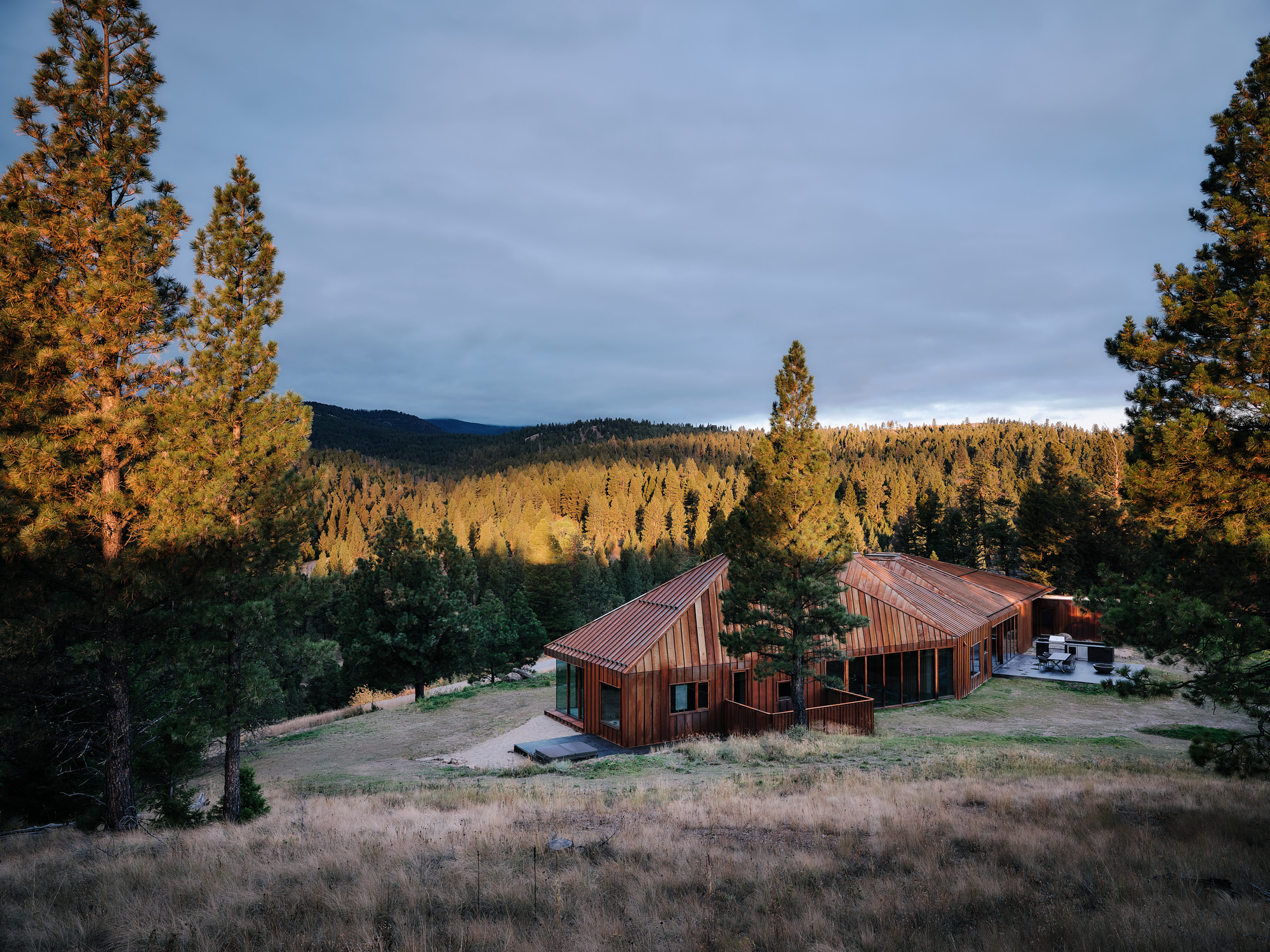 Elevated view of the long metal-clad house with sloping roofs, lit by golden light on the surrounding pine ridge at dusk