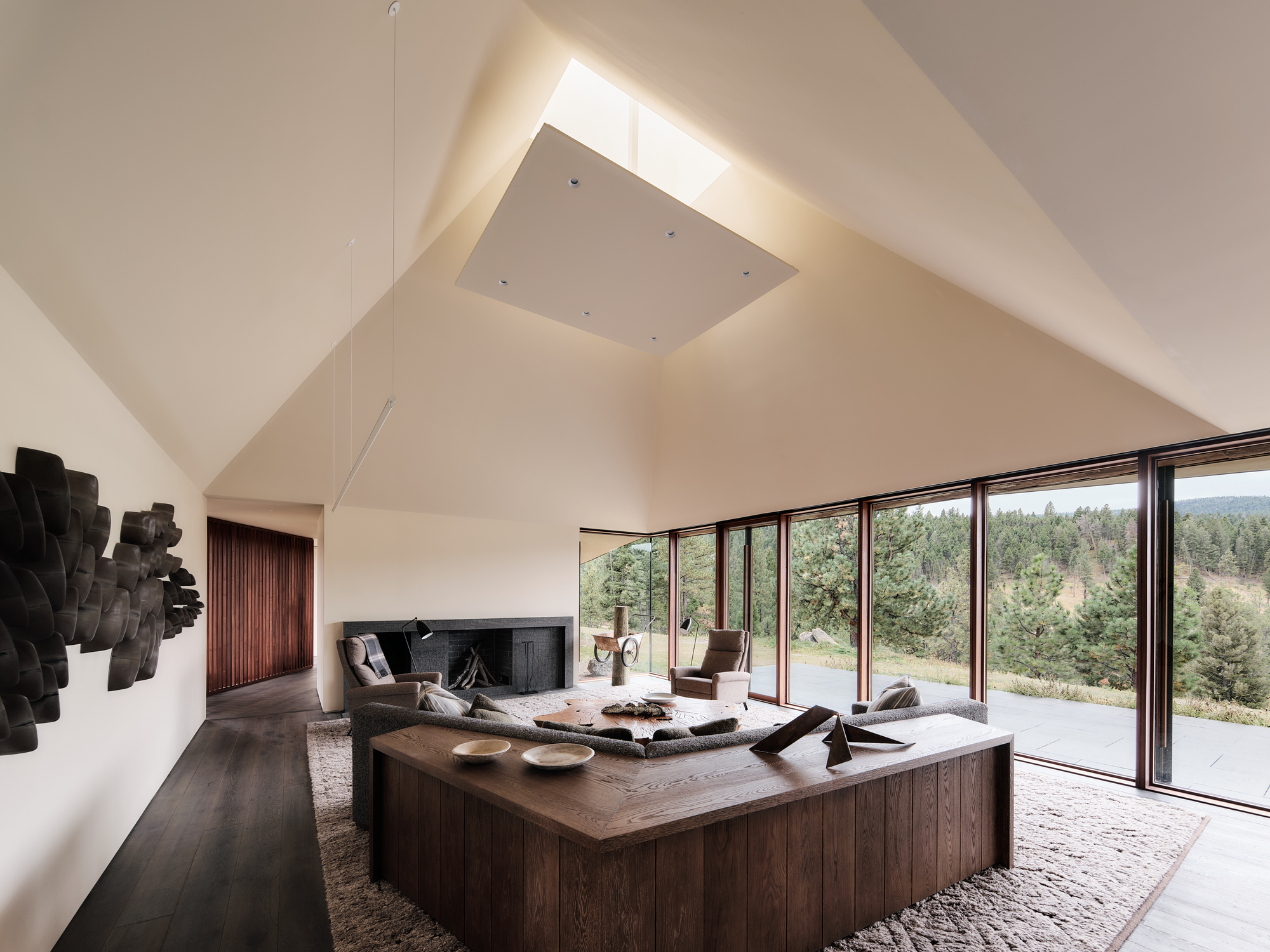Living room with a pitched white ceiling, square skylight and a wood-clad sofa base facing a glass wall onto pines