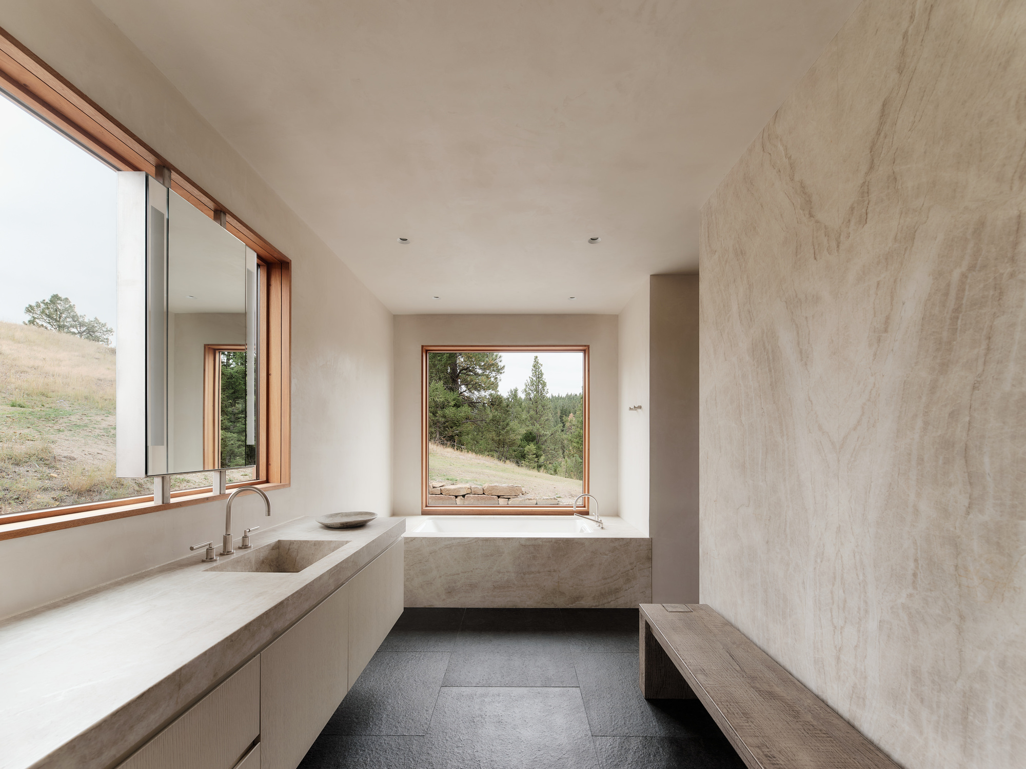 Bathroom with a stone-slab vanity, built-in tub and timber-framed windows opening onto a grassy hillside and pines