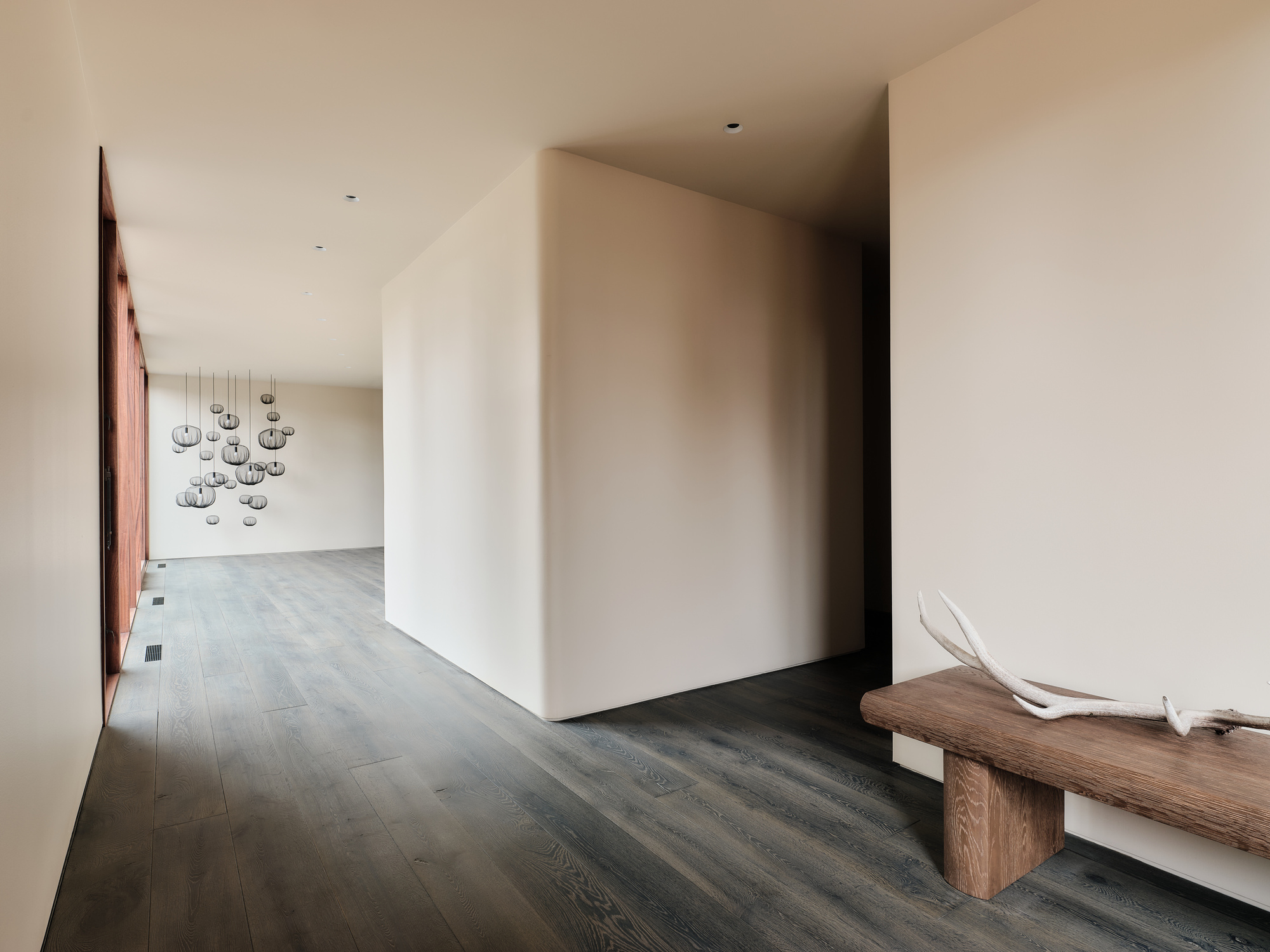Hallway with dark oak floors, a plaster-walled volume, a wooden bench with antlers and a cluster of pendant lamps in the distance