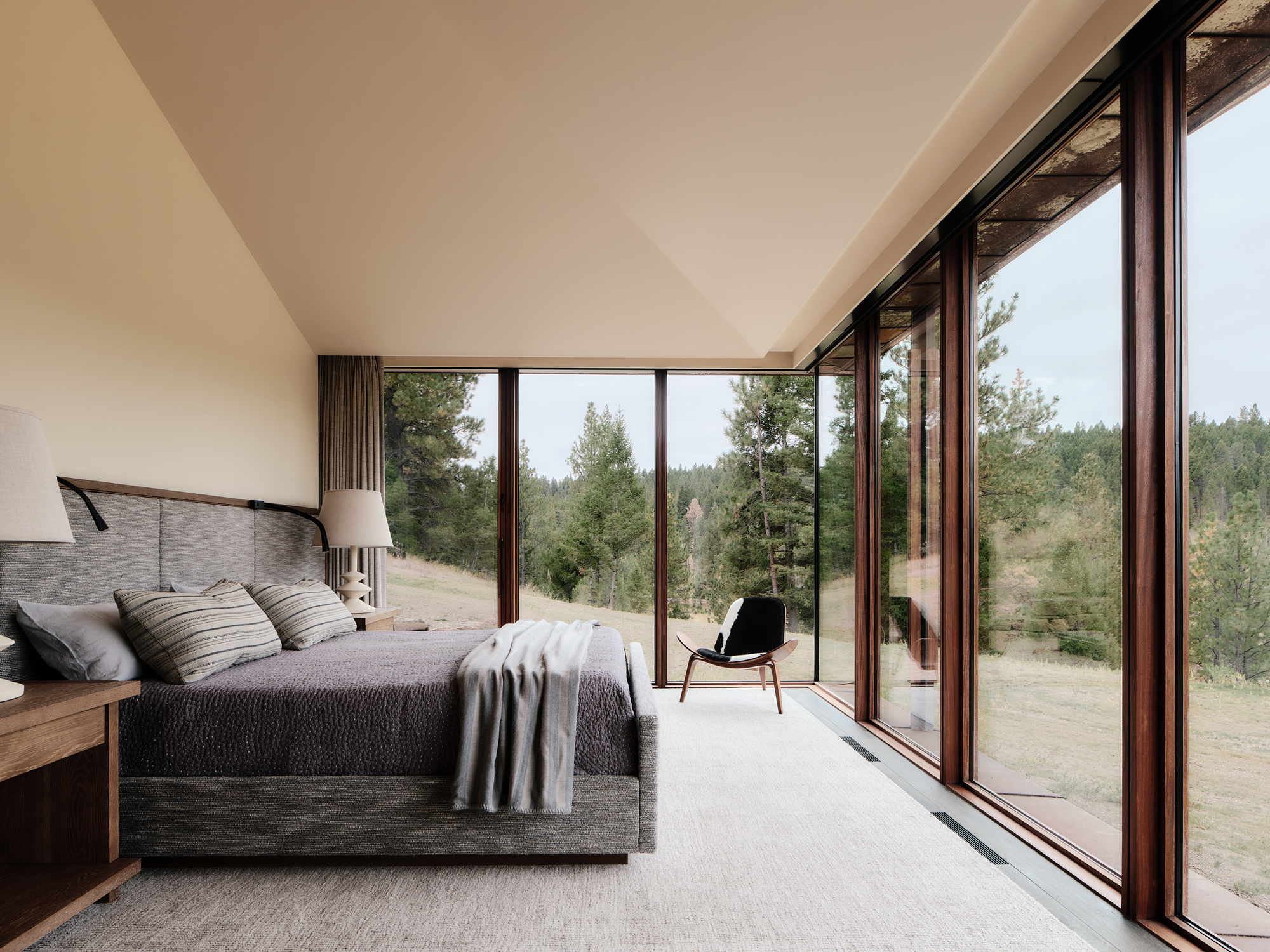 Bedroom corner with floor-to-ceiling walnut-framed glazing, an upholstered bed and a lounge chair facing a pine-covered hillside in overcast light