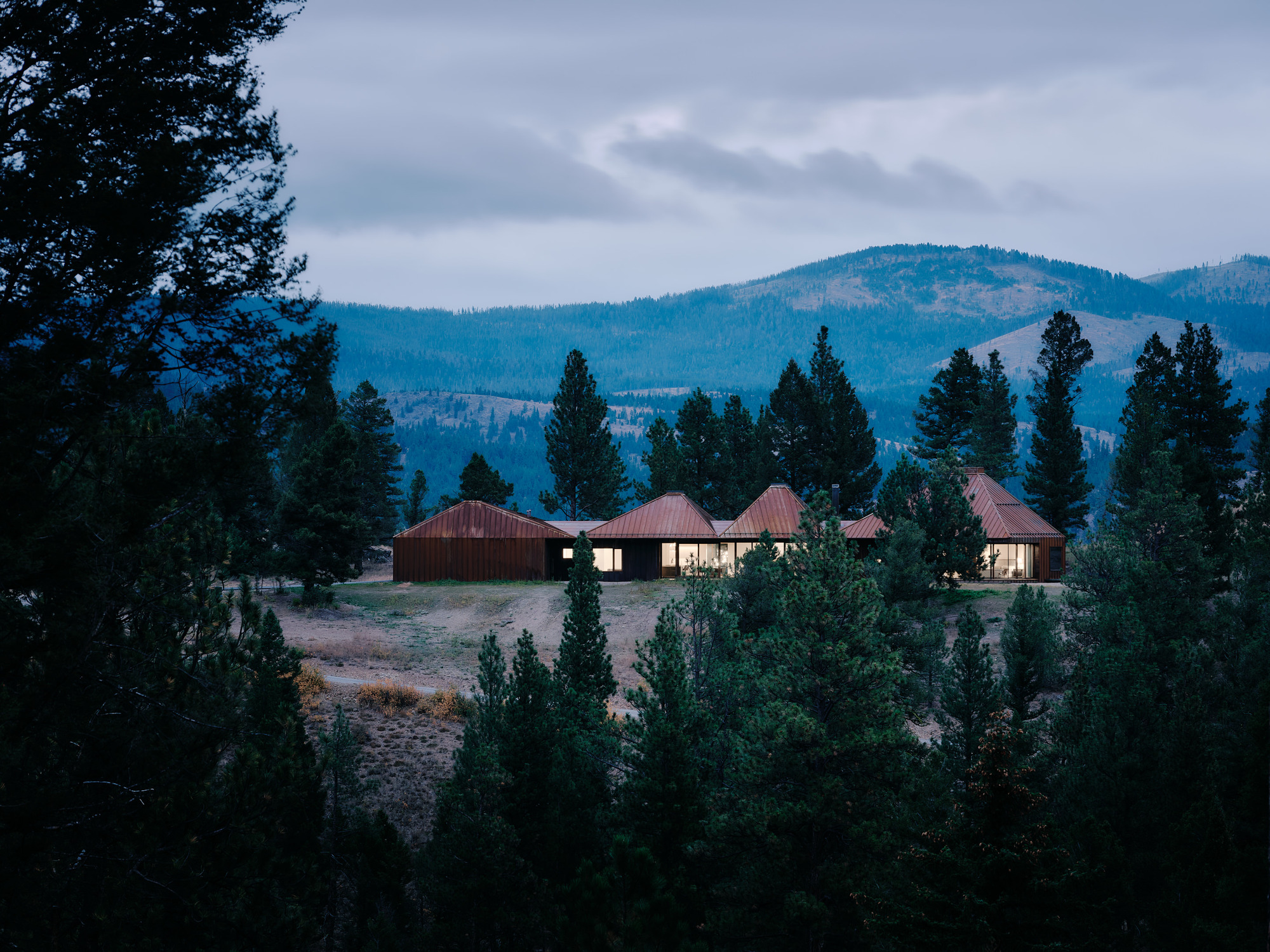 Low house with several pitched copper-toned roofs and lit windows, seen through dark pine trees at dusk with mountains behind