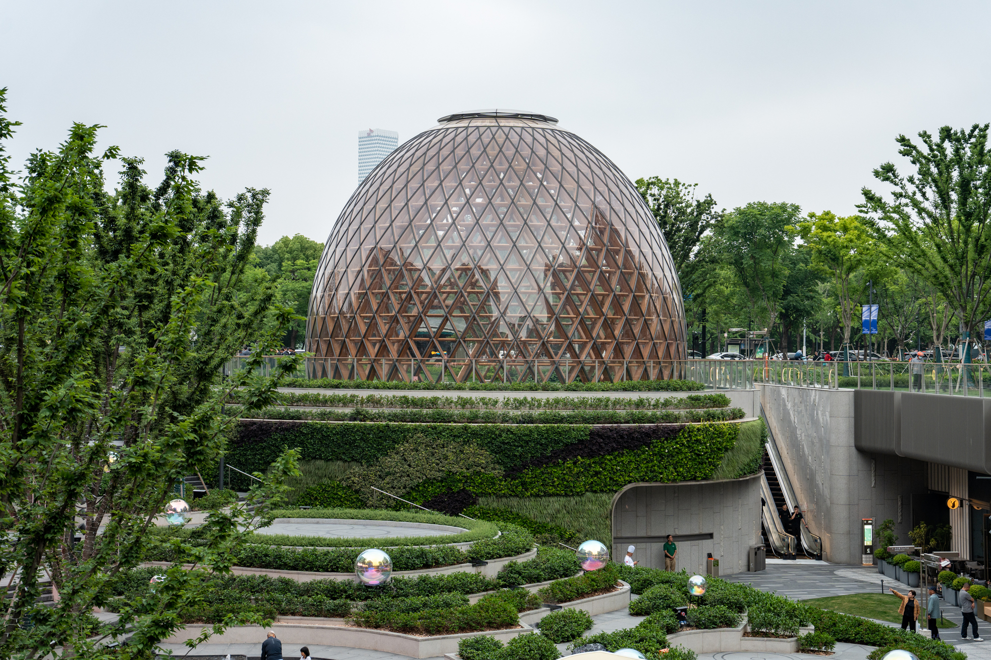 Glazed timber lattice dome above stepped planted terraces, with shoppers near an escalator under an overcast sky