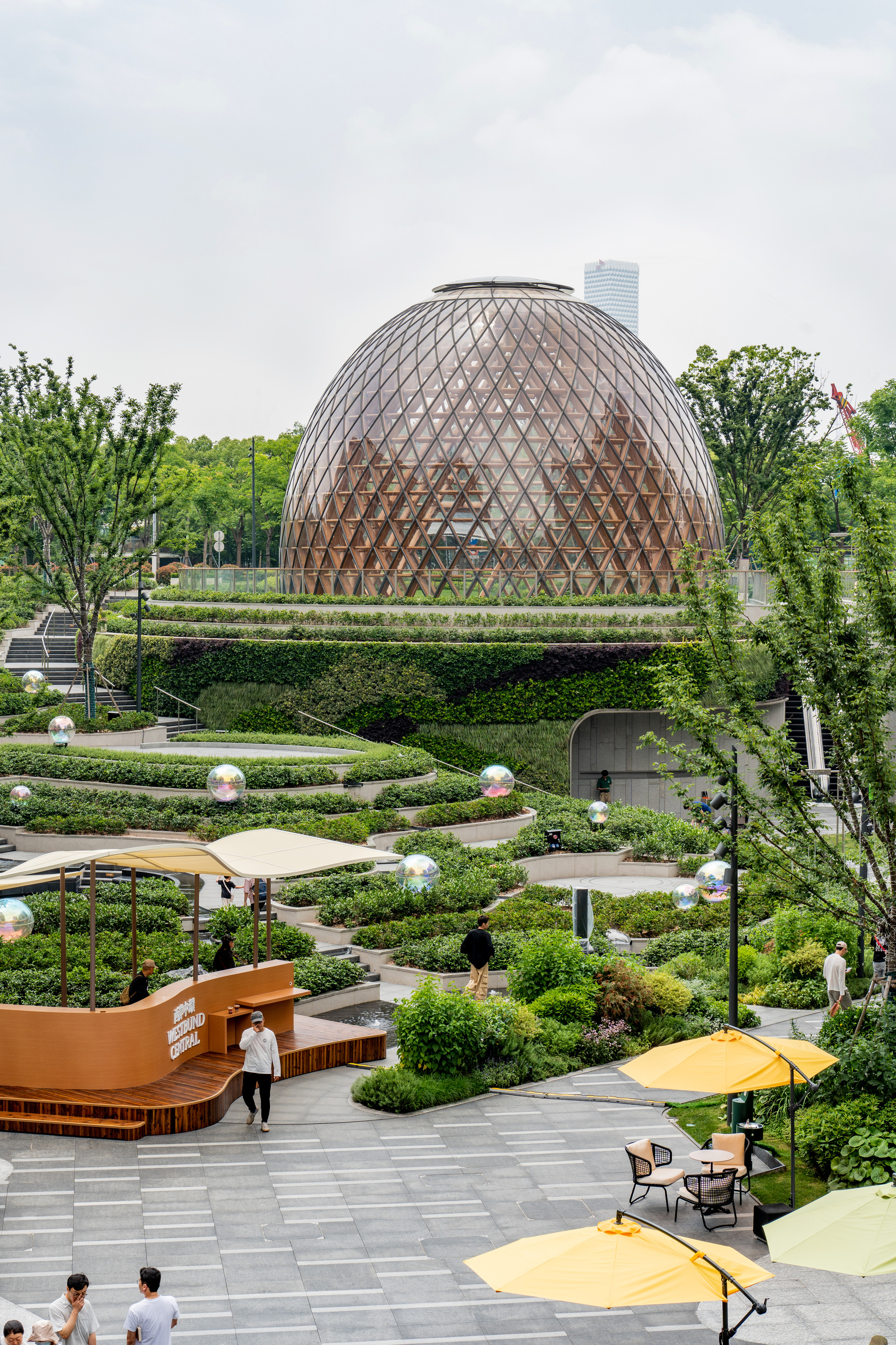 Timber-framed dome rising behind layered planting beds, yellow umbrellas and a curved wooden kiosk, with people crossing the paved plaza