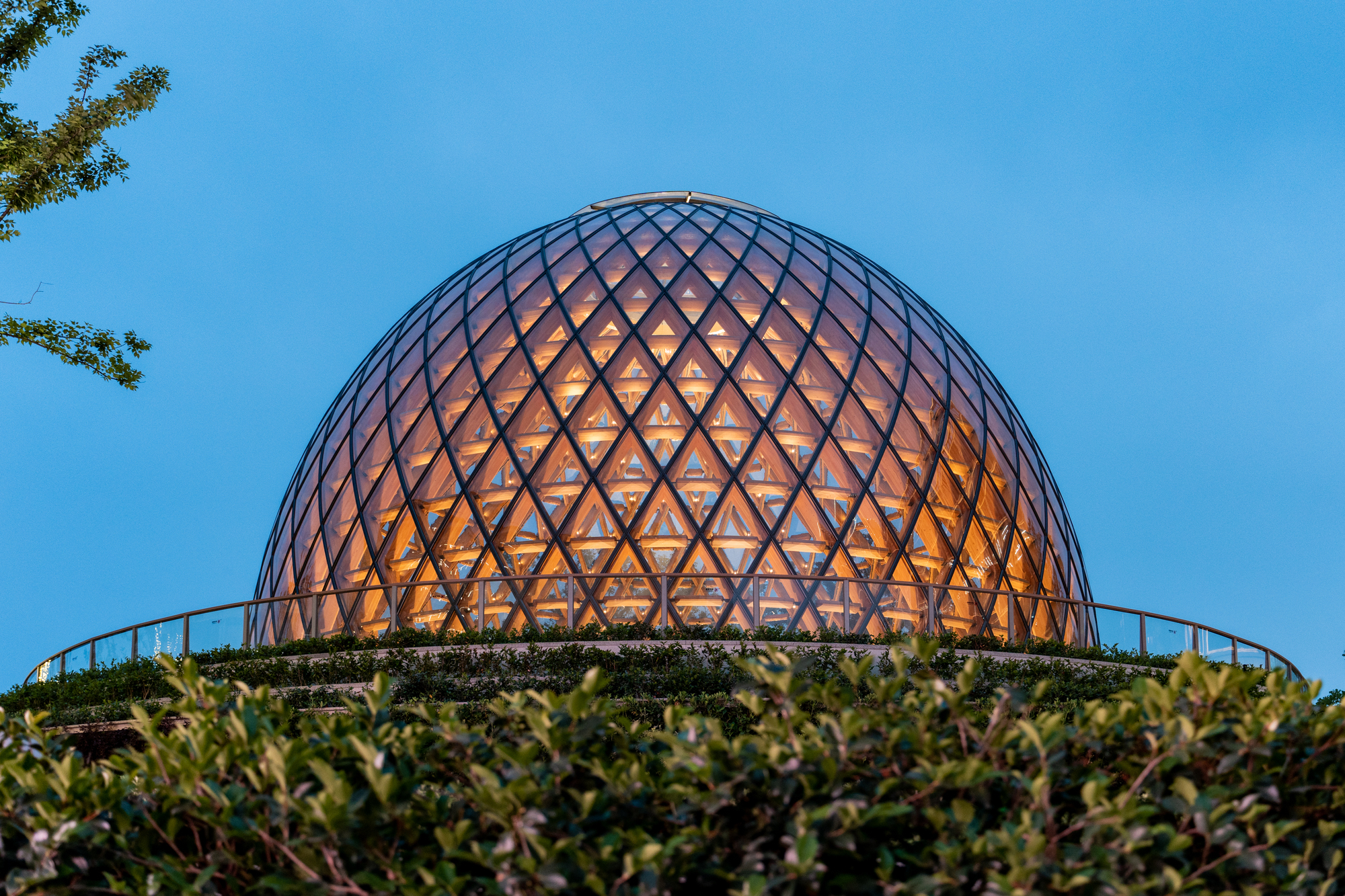 Dome glowing with warm interior light at dusk above a hedge and curved glass balustrade under a deep blue sky