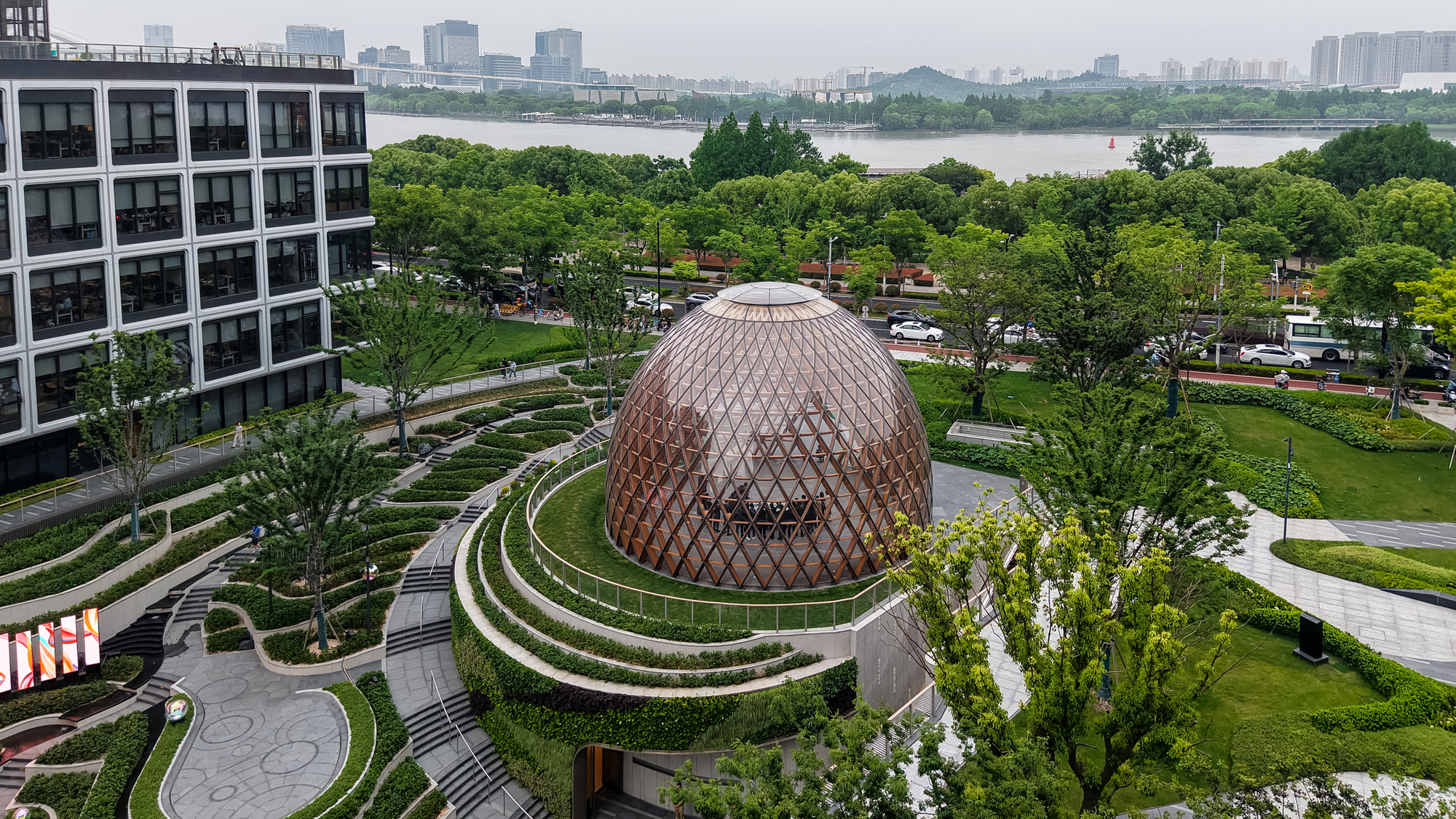 Aerial view of the egg-shaped timber lattice dome ringed by terraced hedges, with a river and hazy skyline beyond