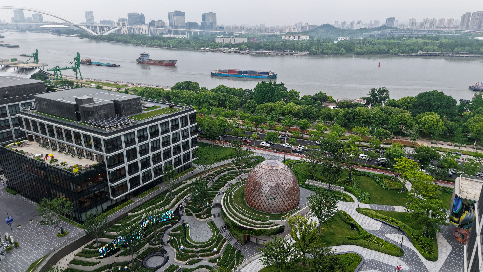 Drone view of the domed pavilion in a terraced garden between office blocks, with cargo barges on the river behind