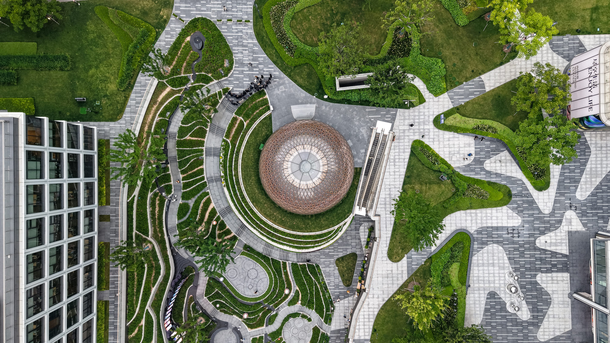 Top-down view of the lattice dome encircled by curving planted terraces and a pavement of irregular paving patterns