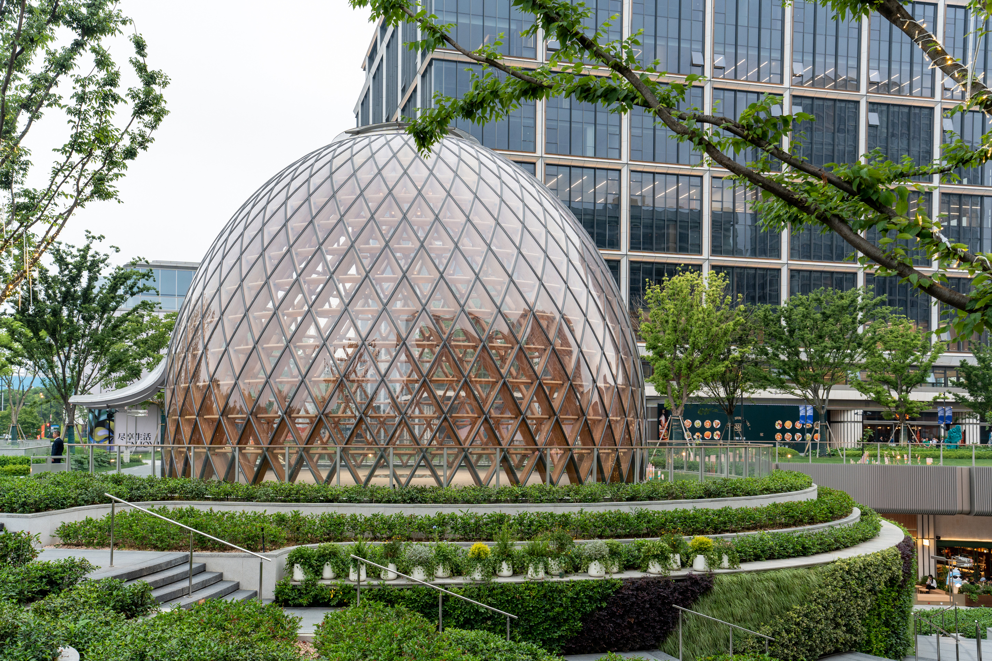 Diamond-gridded glass dome above layered planted terraces, with a leafy branch framing the glass office tower behind