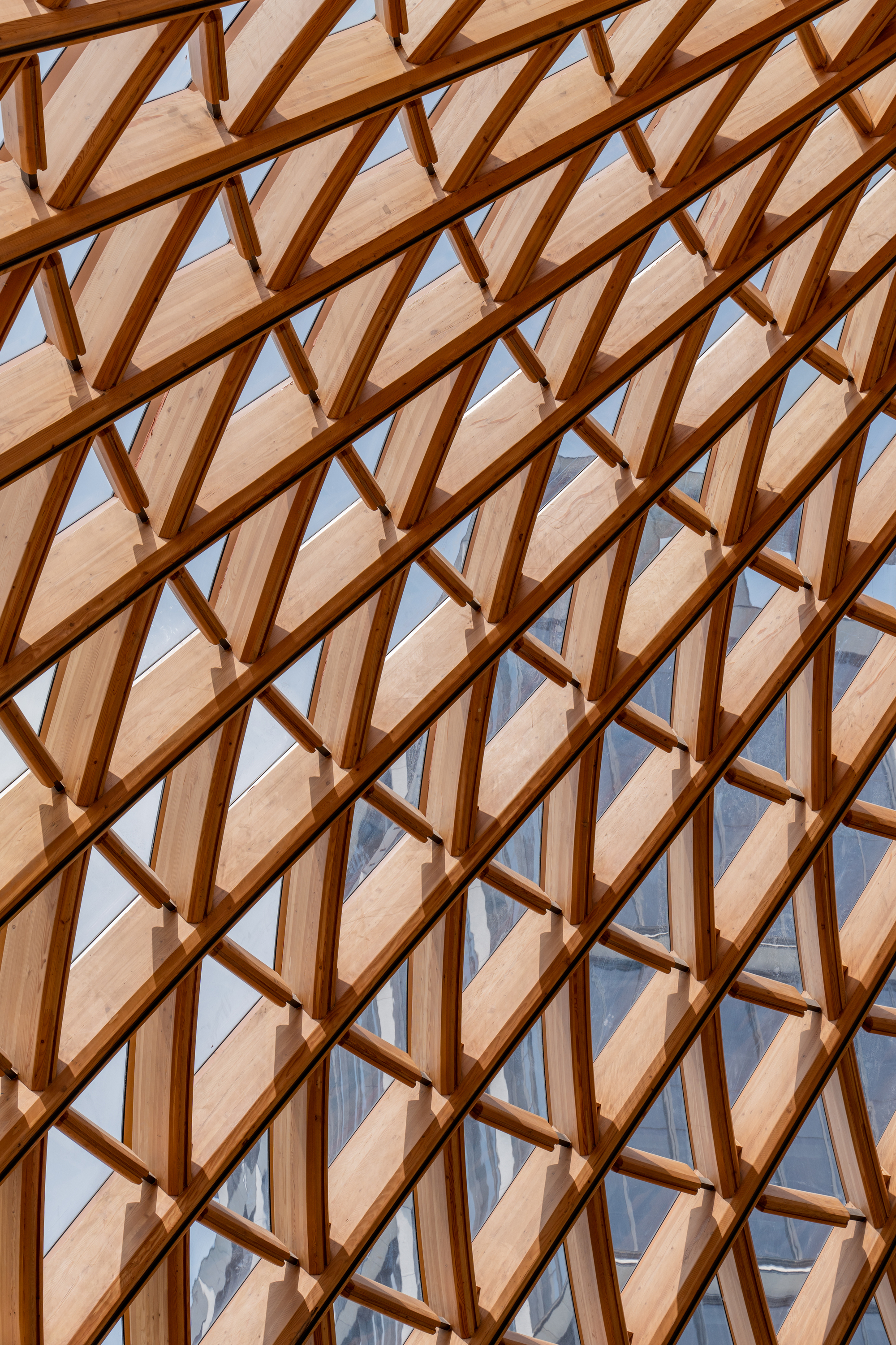 Tight view of overlapping timber lattice members and glass panels, with reflected sky and neighboring buildings in the panes