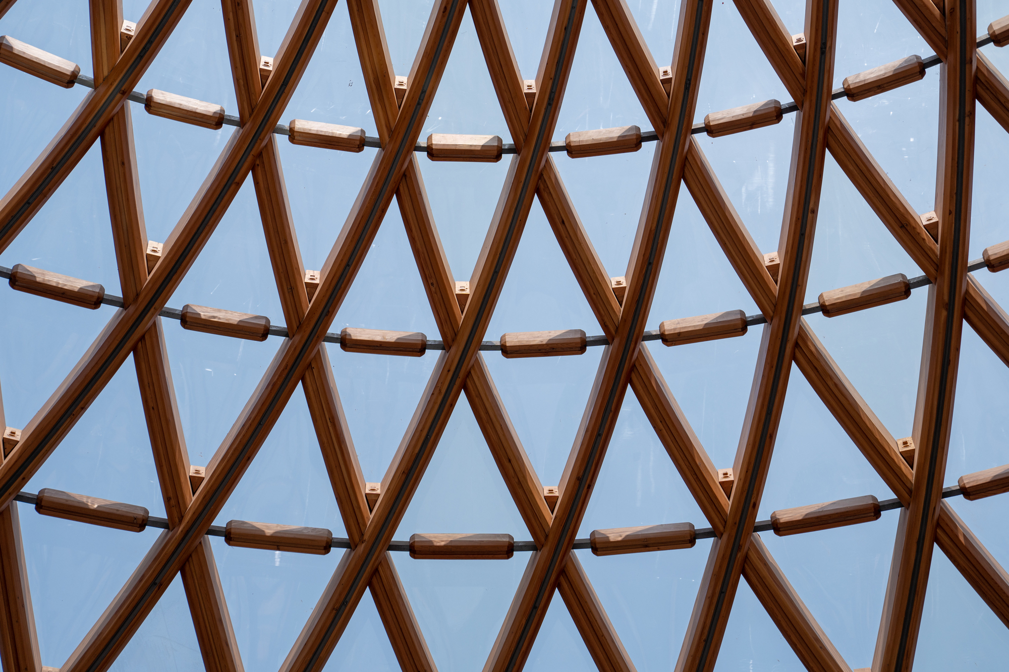 Looking up through a triangulated timber grid with small wooden blocks spaced along the glazing, against a pale blue sky