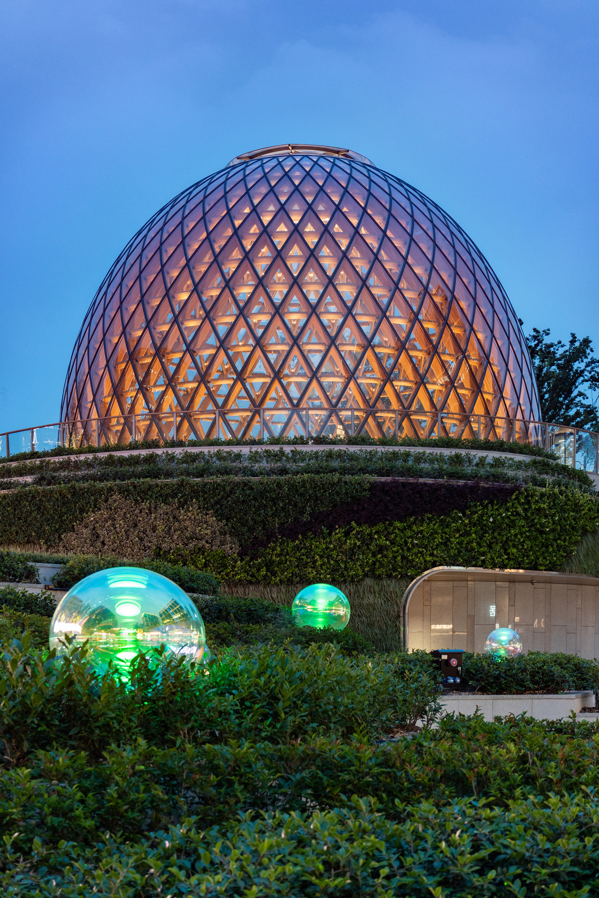 Egg-shaped timber and glass dome glowing at dusk above terraced hedges and lit glass spheres