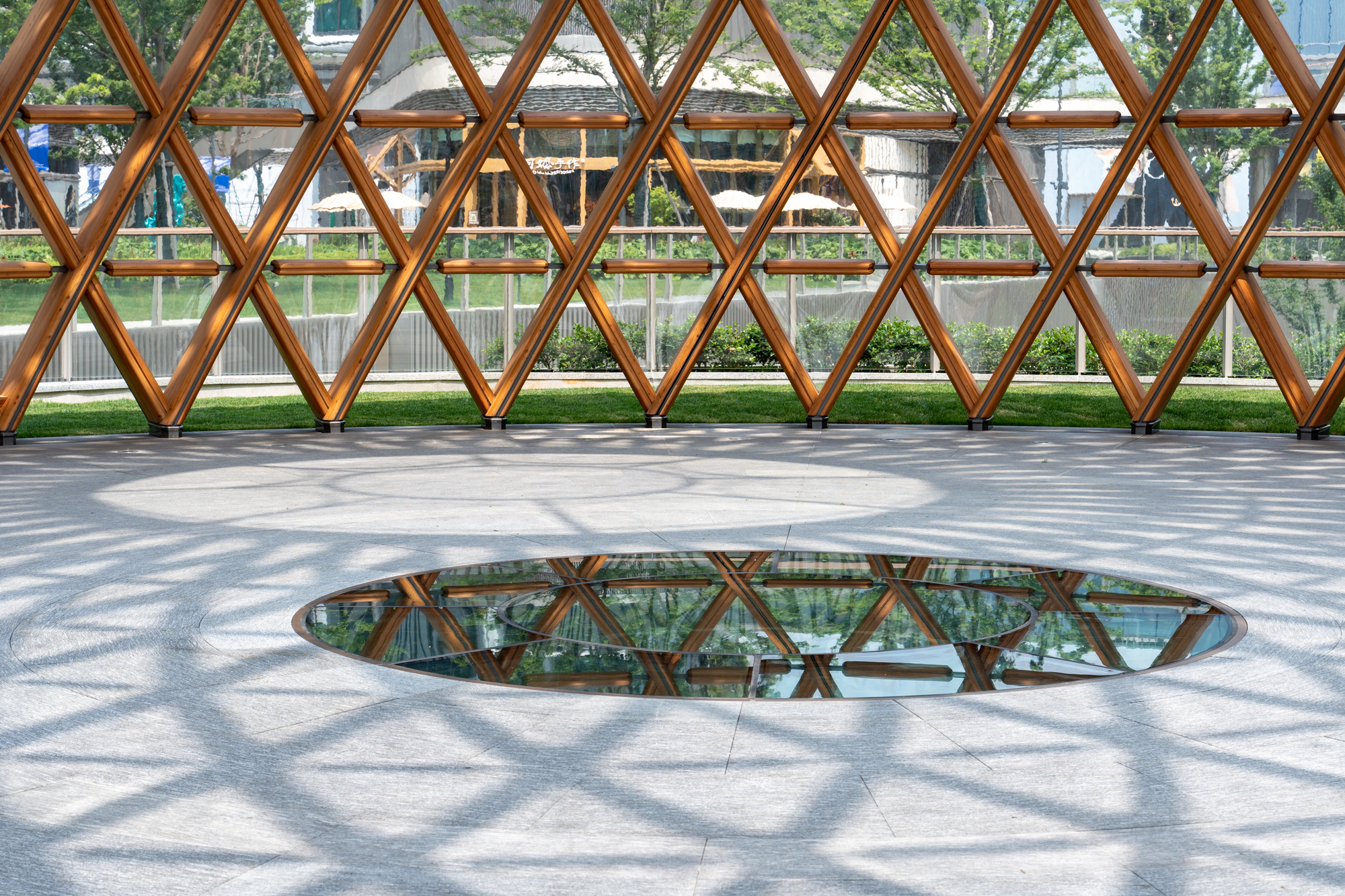 Diamond lattice of timber members above a paved floor with a round glass inset, casting latticed shadows in bright sun