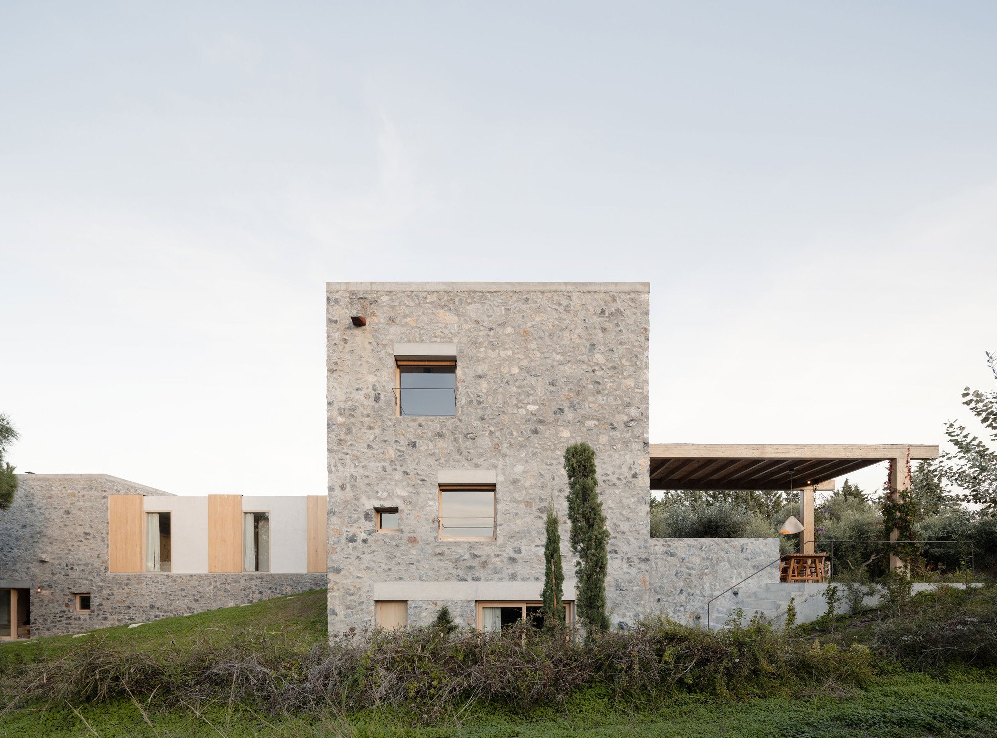 Stone tower volume beside a low wing with timber shutters and a pergola roof, seen across overgrown grass under pale evening sky