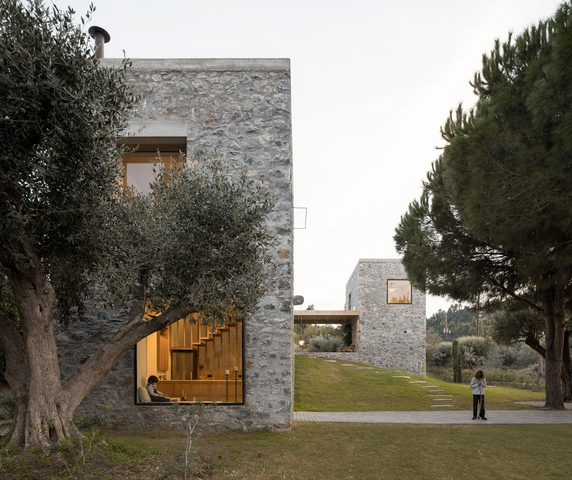 Stone facade with a large corner window framing a timber stair, an olive tree in front and a child at right