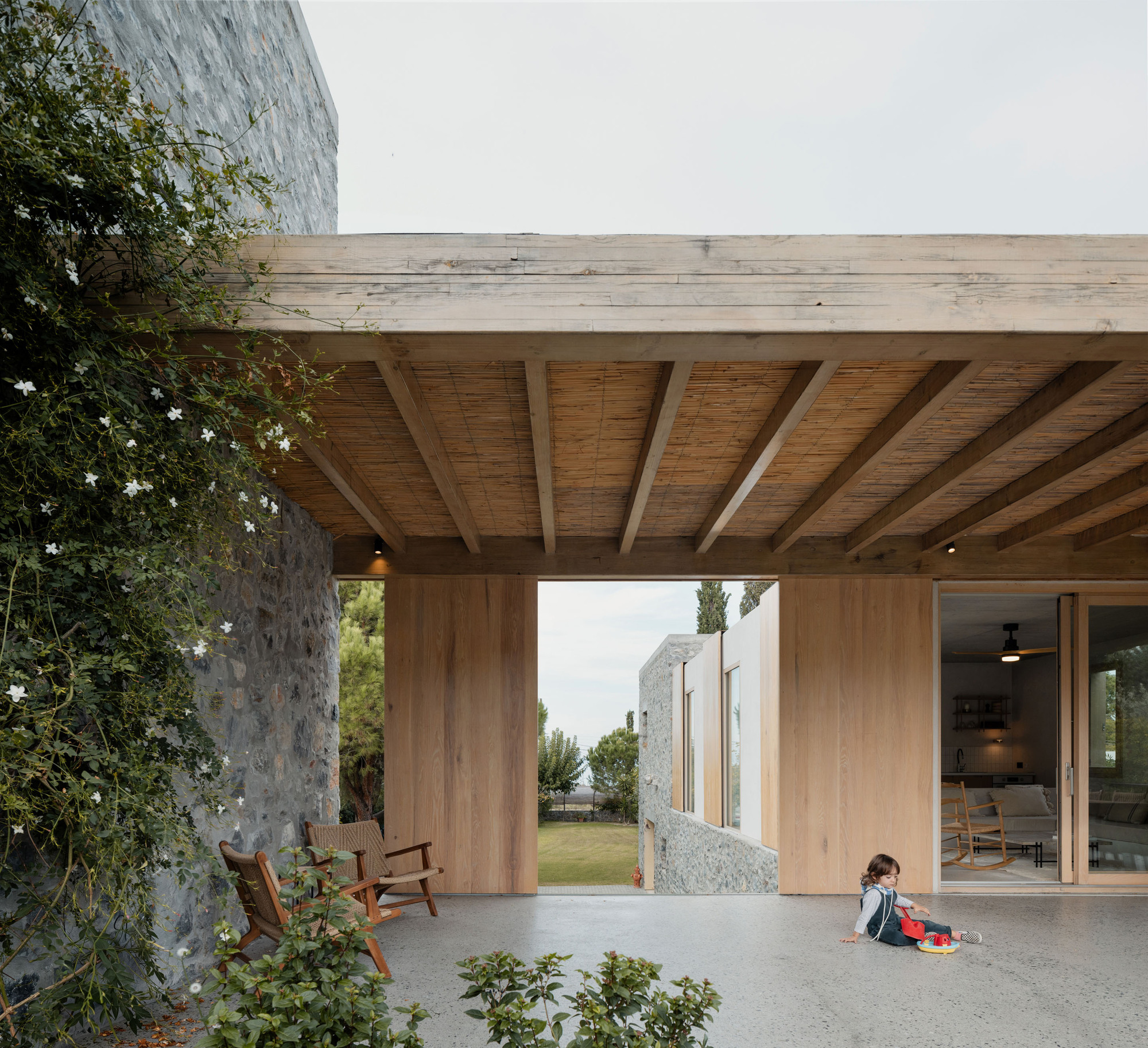 Covered porch with timber beams and reed ceiling panels, a jasmine-clad stone wall and a child playing on the concrete floor