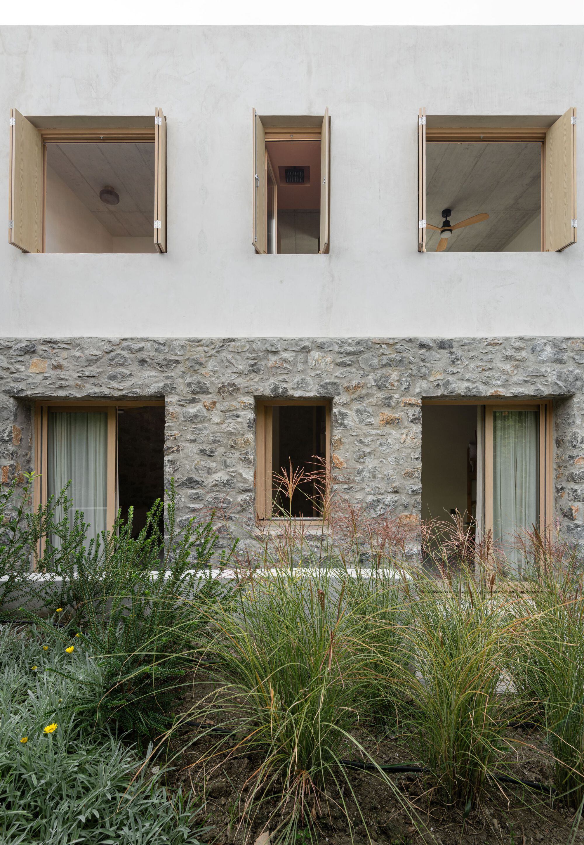 Facade with open timber shutters on a white plaster upper floor above a stone base, with grasses in the foreground